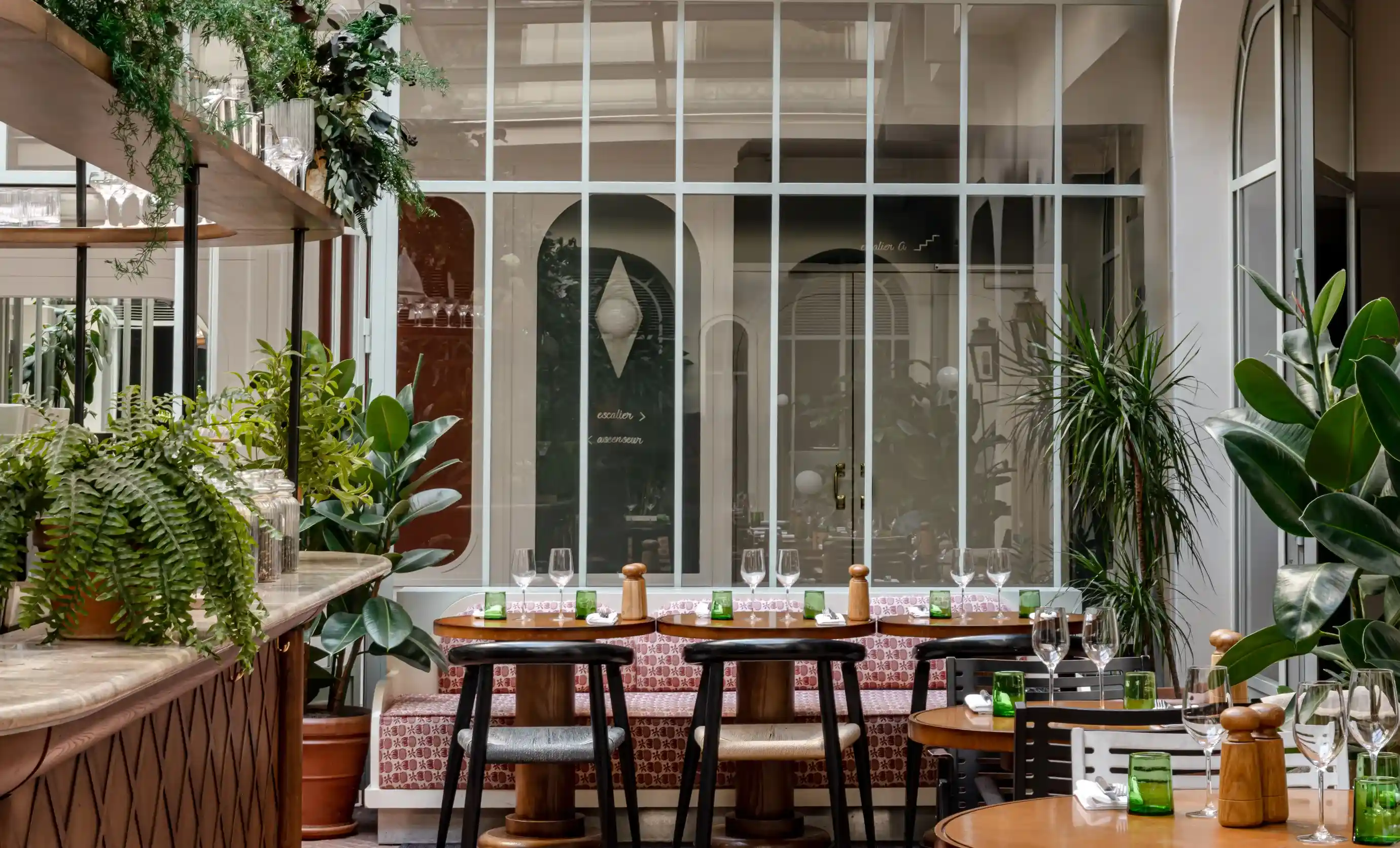 Cozy restaurant seating with wooden tables, chairs, green plants, and glassware on tables.
