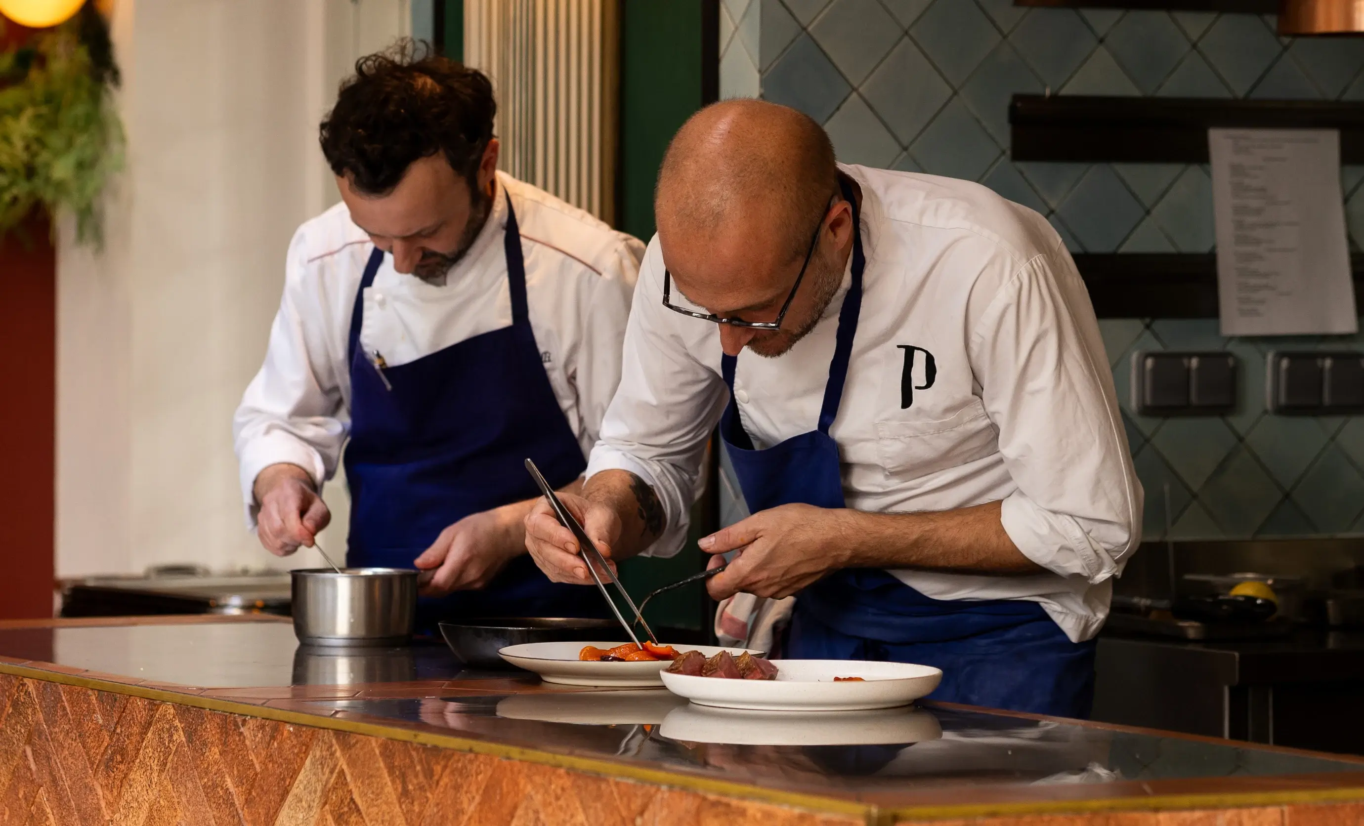 Two chefs in white shirts and blue aprons carefully plating food in a professional kitchen.