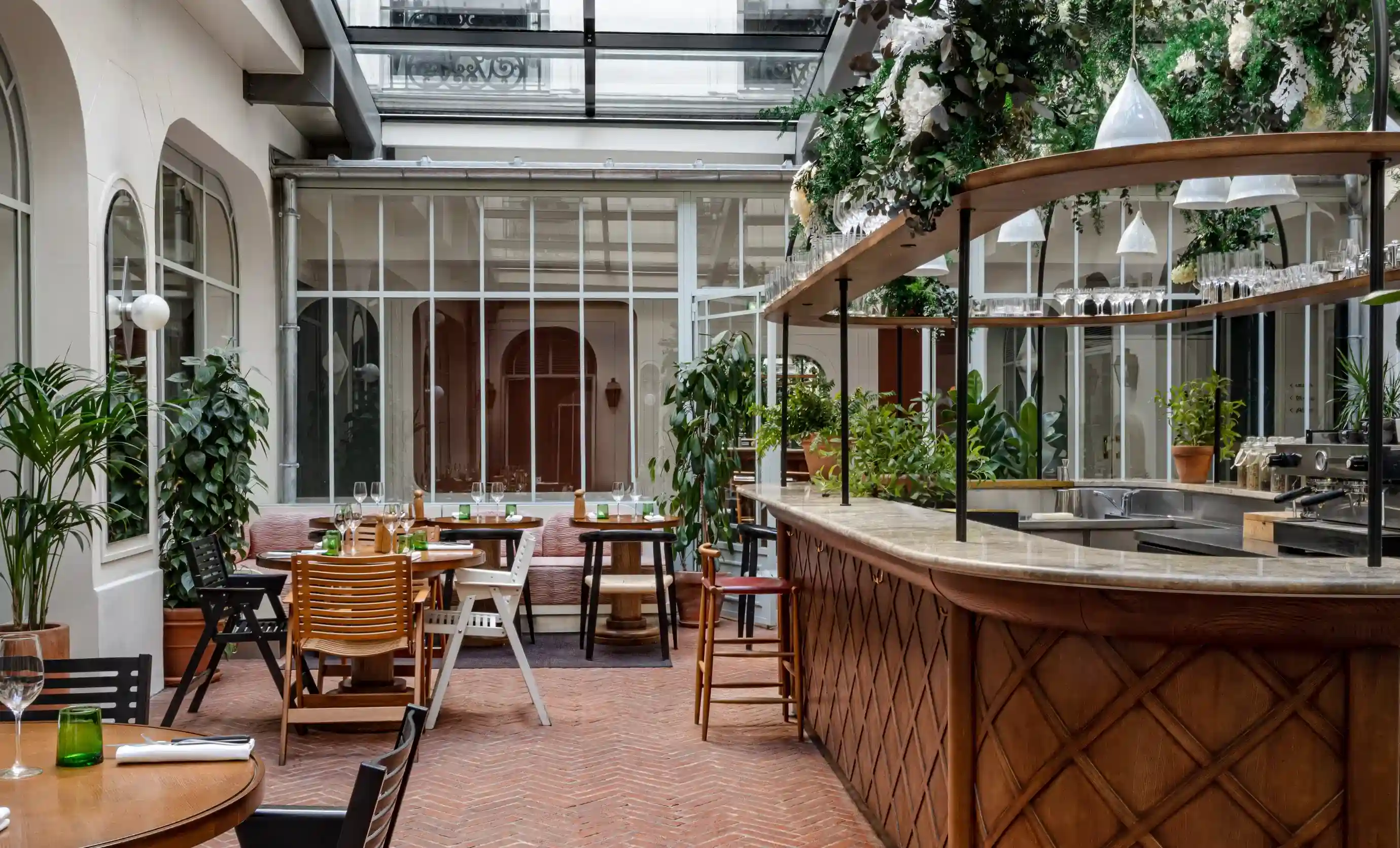 Indoor restaurant with wooden tables, mixed chairs, plants, and a curved bar under glass ceiling.