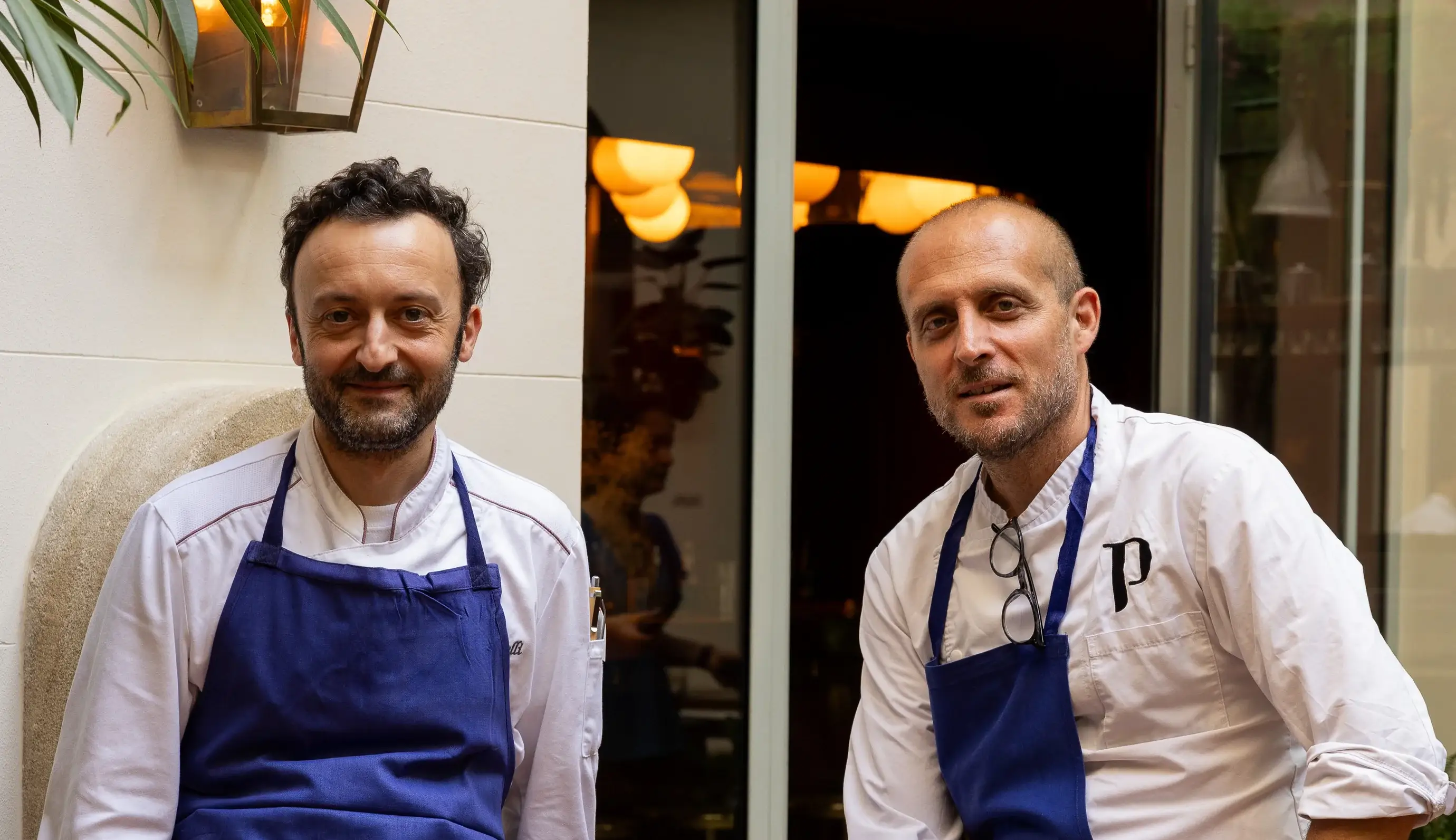 Two male chefs wearing blue aprons and white shirts standing in front of a glass door.