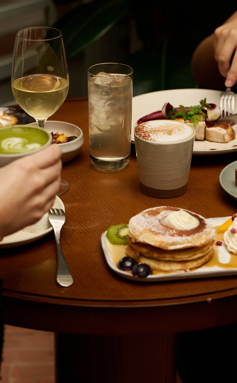 Brunch table with pancakes, cappuccino, green tea latte, wine, and a plate with meat and garnish.