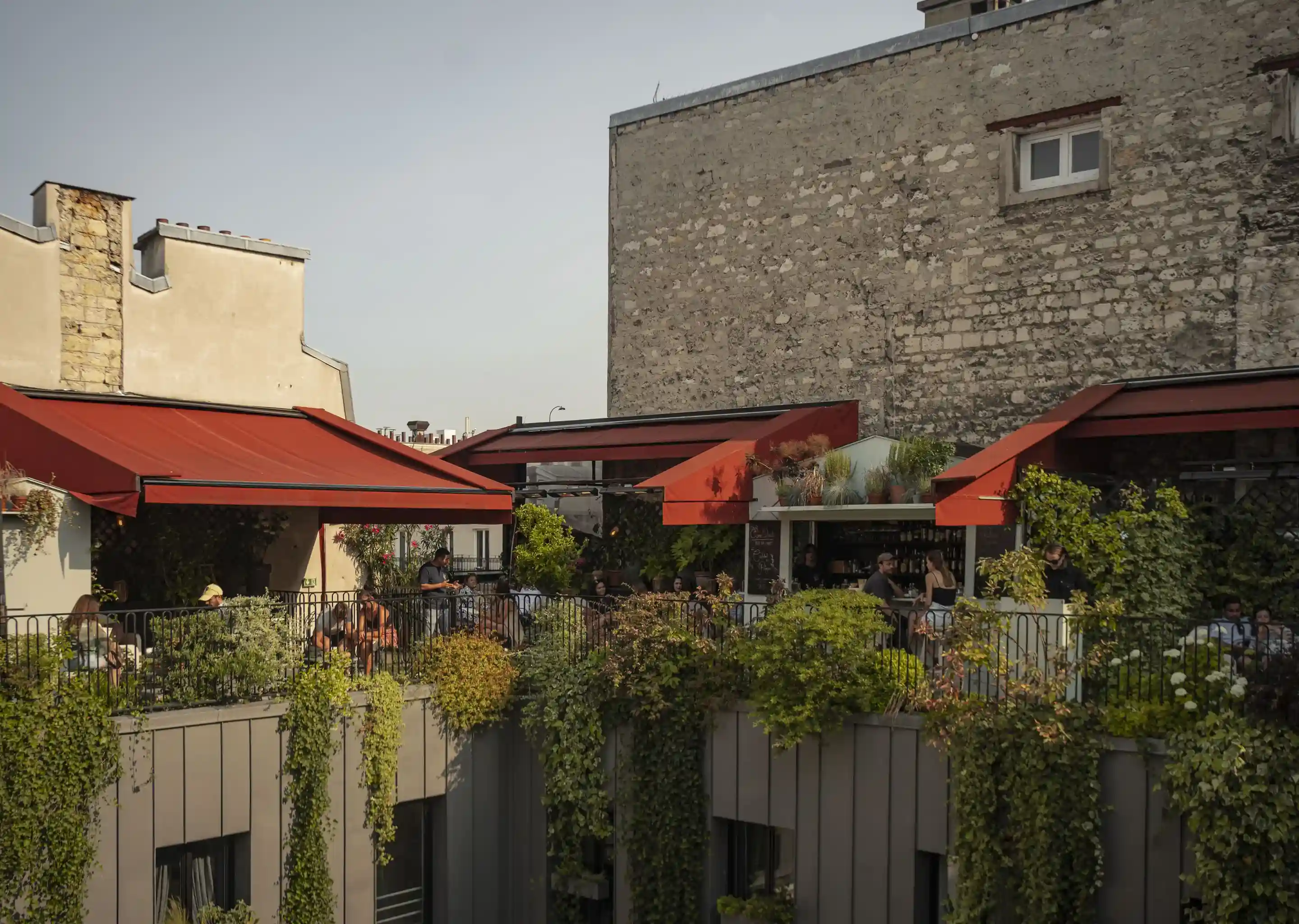 Outdoor rooftop bar with red awnings, green plants, and people seated socializing under evening light.