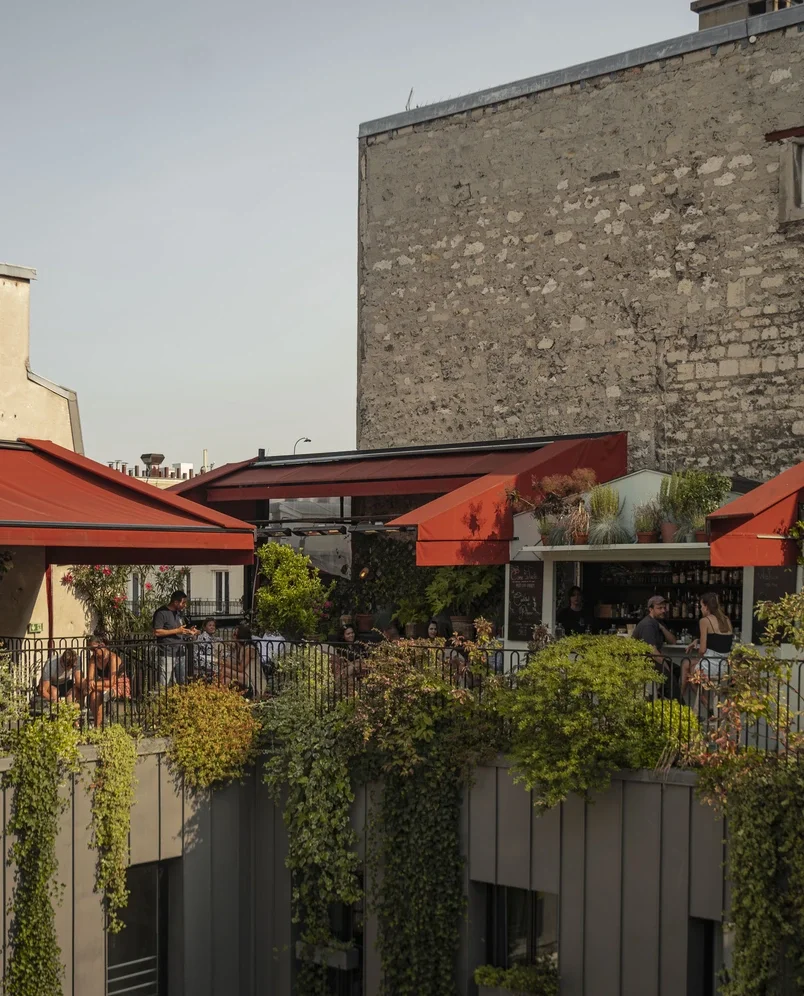 Outdoor rooftop bar terrace with people sitting under orange awnings and green plants cascading down.
