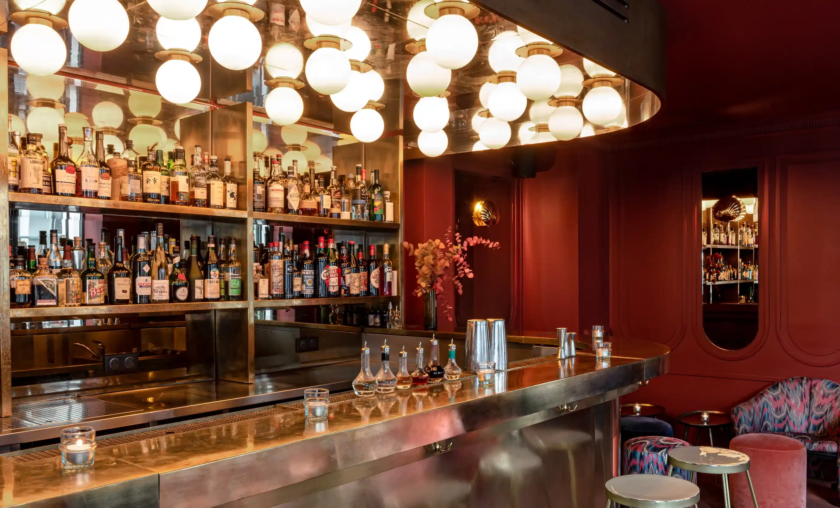 Bar moderno com balcão de latão, prateleiras de garrafas de bebidas, luzes redondas suspensas e área de estar colorida.