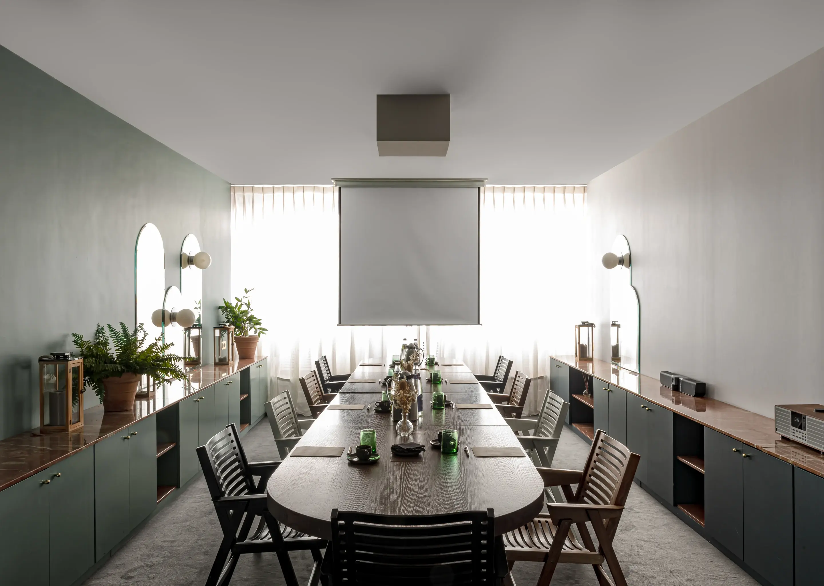 Sala de conferências moderna com longa mesa de madeira, cadeiras, plantas nos armários laterais e uma tela de projetor em branco.