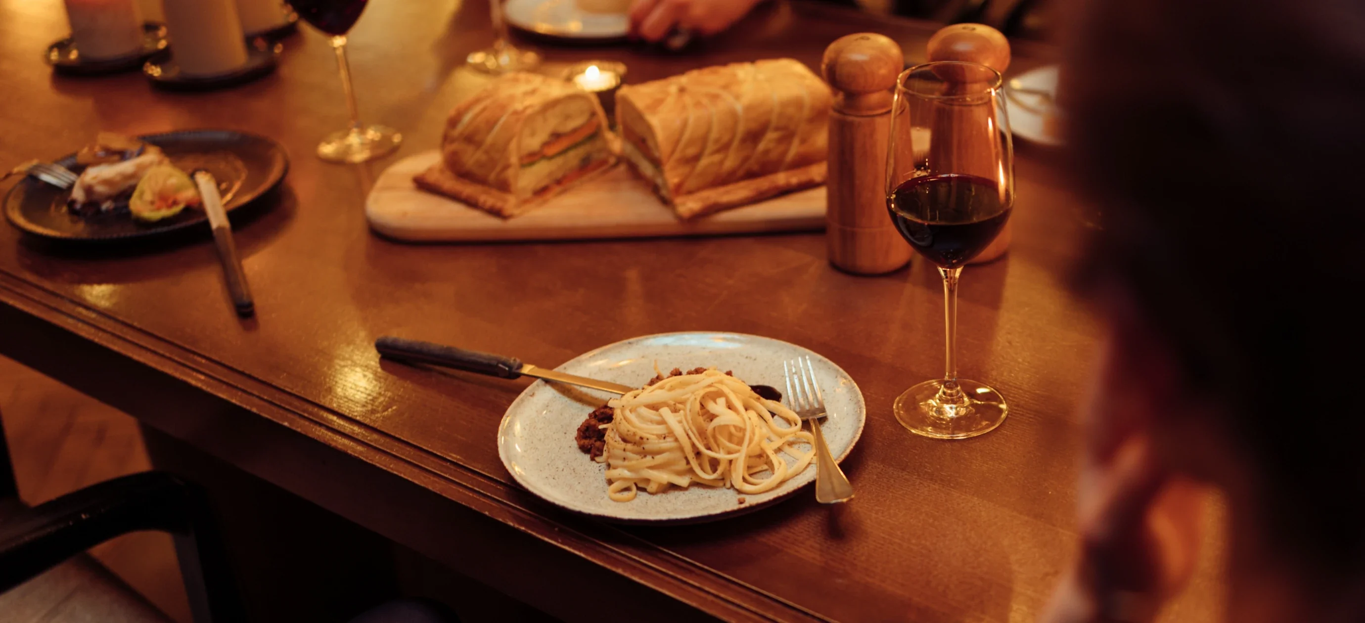 Prato de macarrão com molho, sanduíche na tábua de cortar, copo de vinho tinto e moedores de pimenta na mesa de madeira.