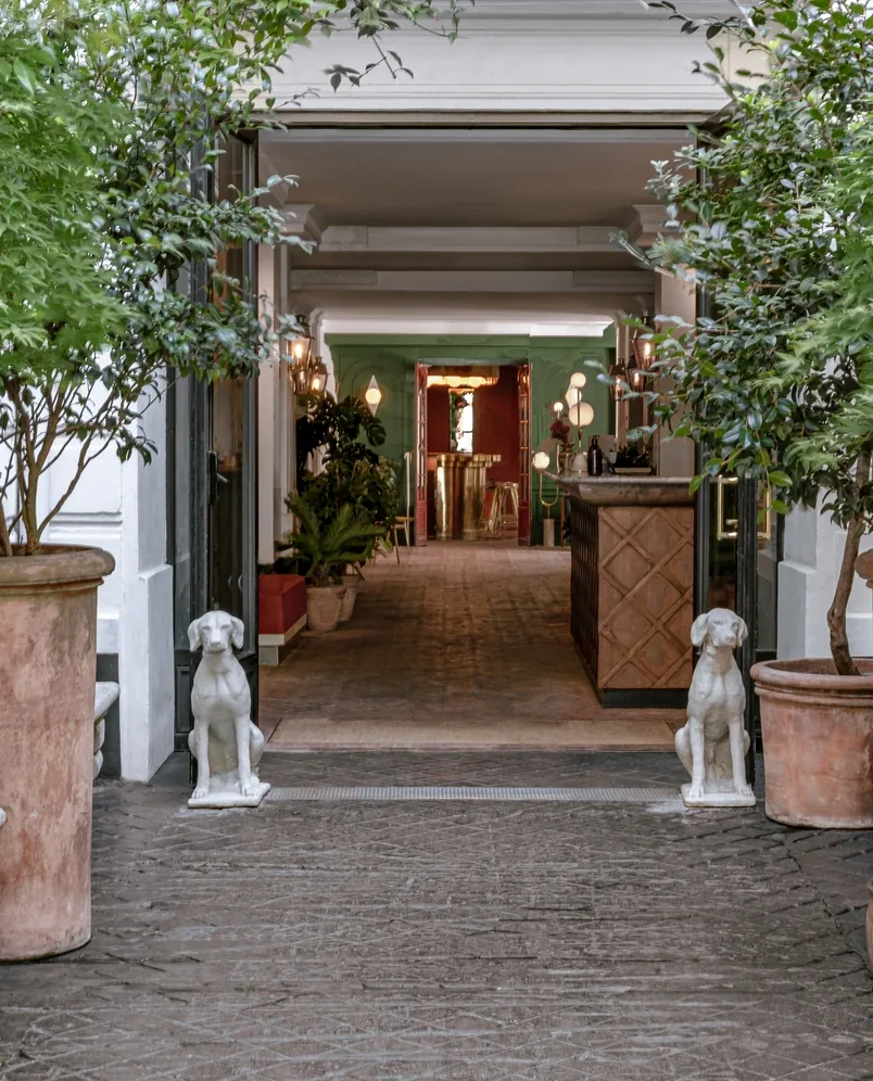 Entrada para um pátio com duas estátuas de cães brancos, grandes vasos de plantas e vista para um elegante corredor interior.