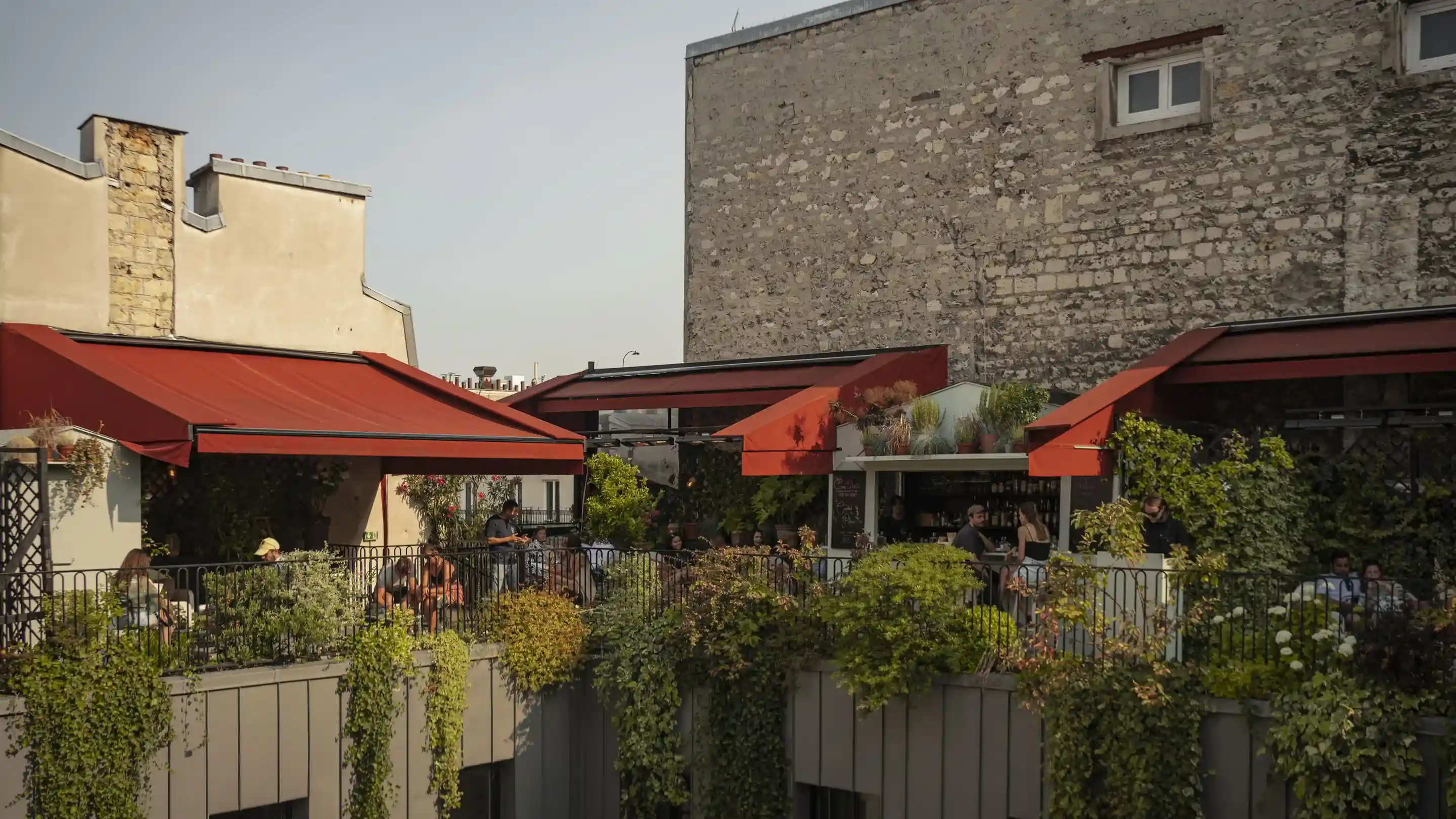 Caffè con terrazza all'aperto con tende rosse, piante verdi e persone sedute ai tavoli sotto la luce del sole.