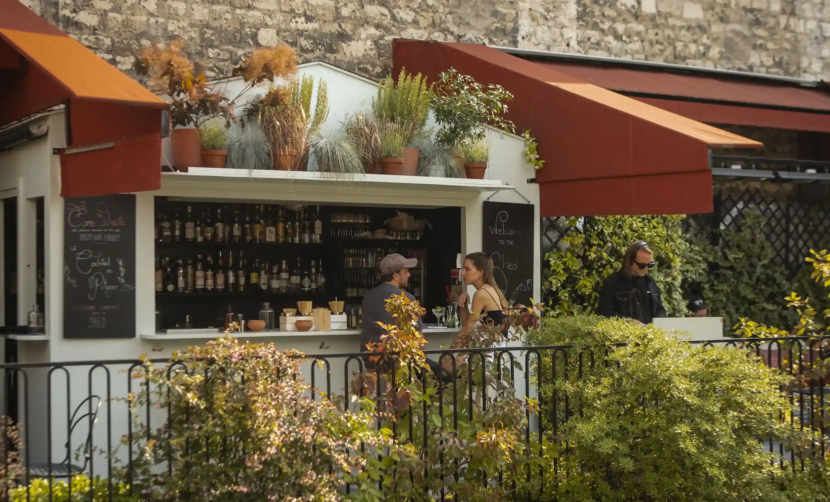 Bar all'aperto con tende rosse, due persone sedute al bancone e un uomo in piedi nelle vicinanze con il verde intorno.