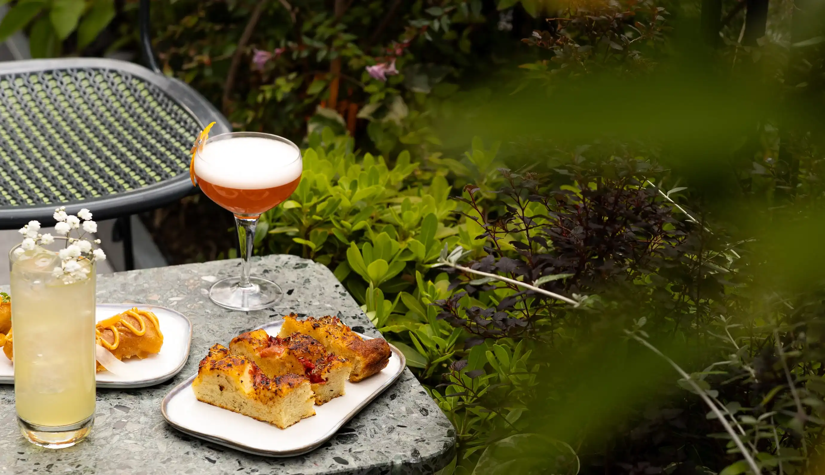 Tavolo con focaccia, pasticcini, una bevanda con guarnizione floreale e un cocktail all'aperto immerso nel verde.
