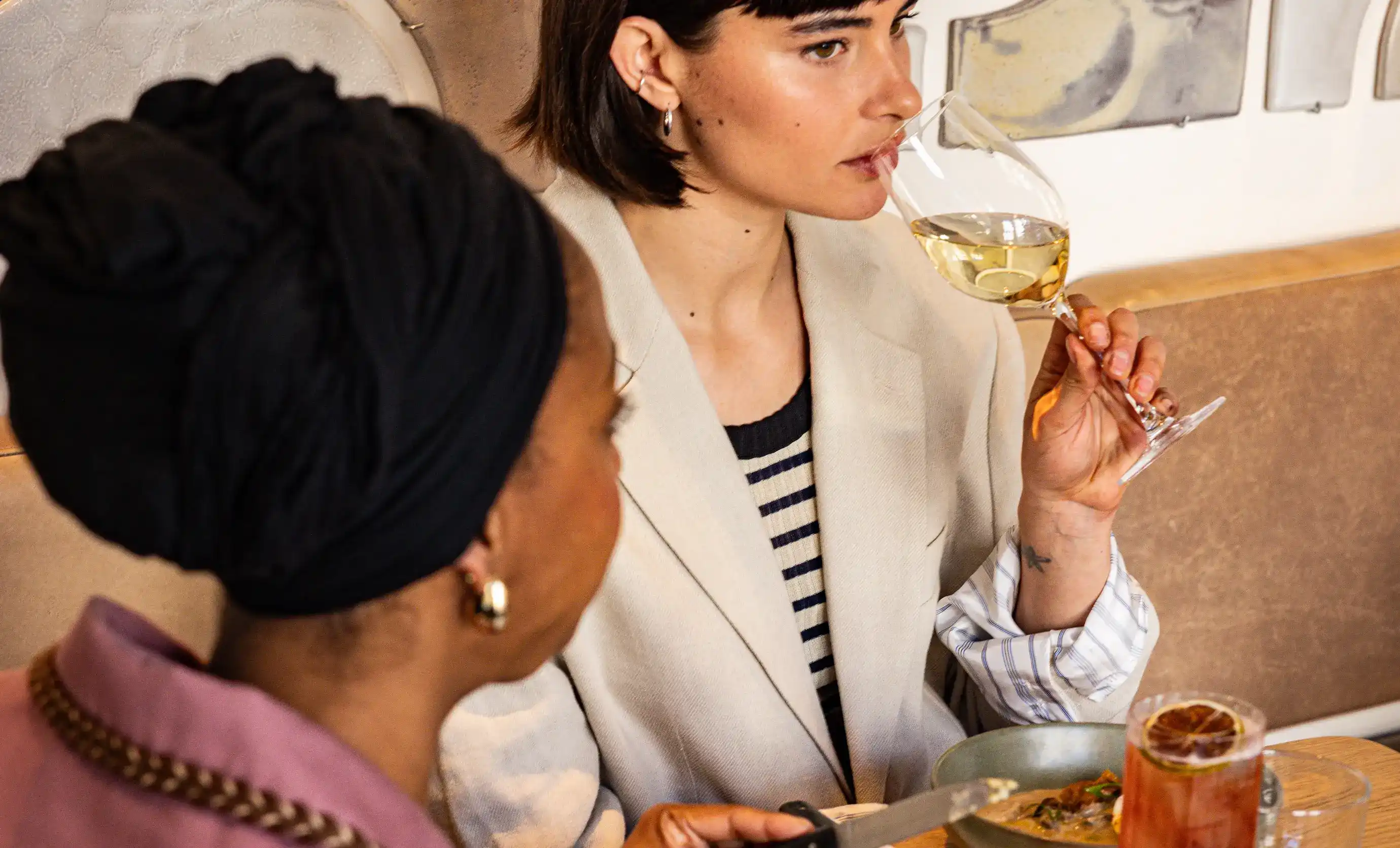 Duas mulheres jantando, uma com um lenço preto e a outra bebendo vinho branco na mesa de um restaurante.