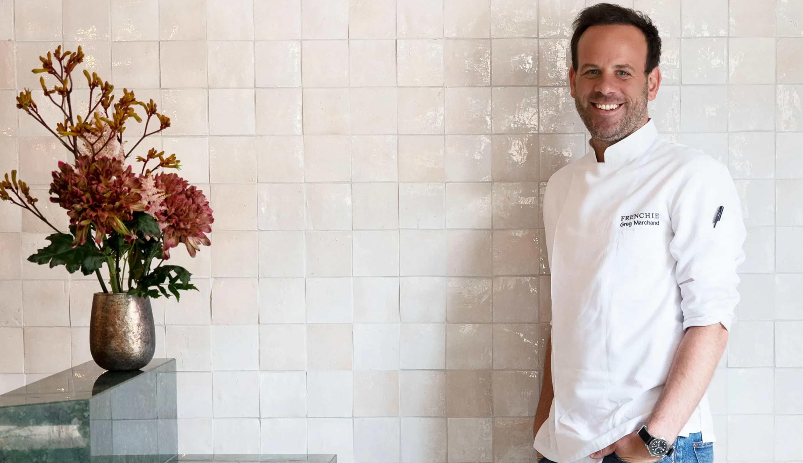 Chef sorridente de jaleco branco ao lado de um vaso de flores contra uma parede de azulejos.