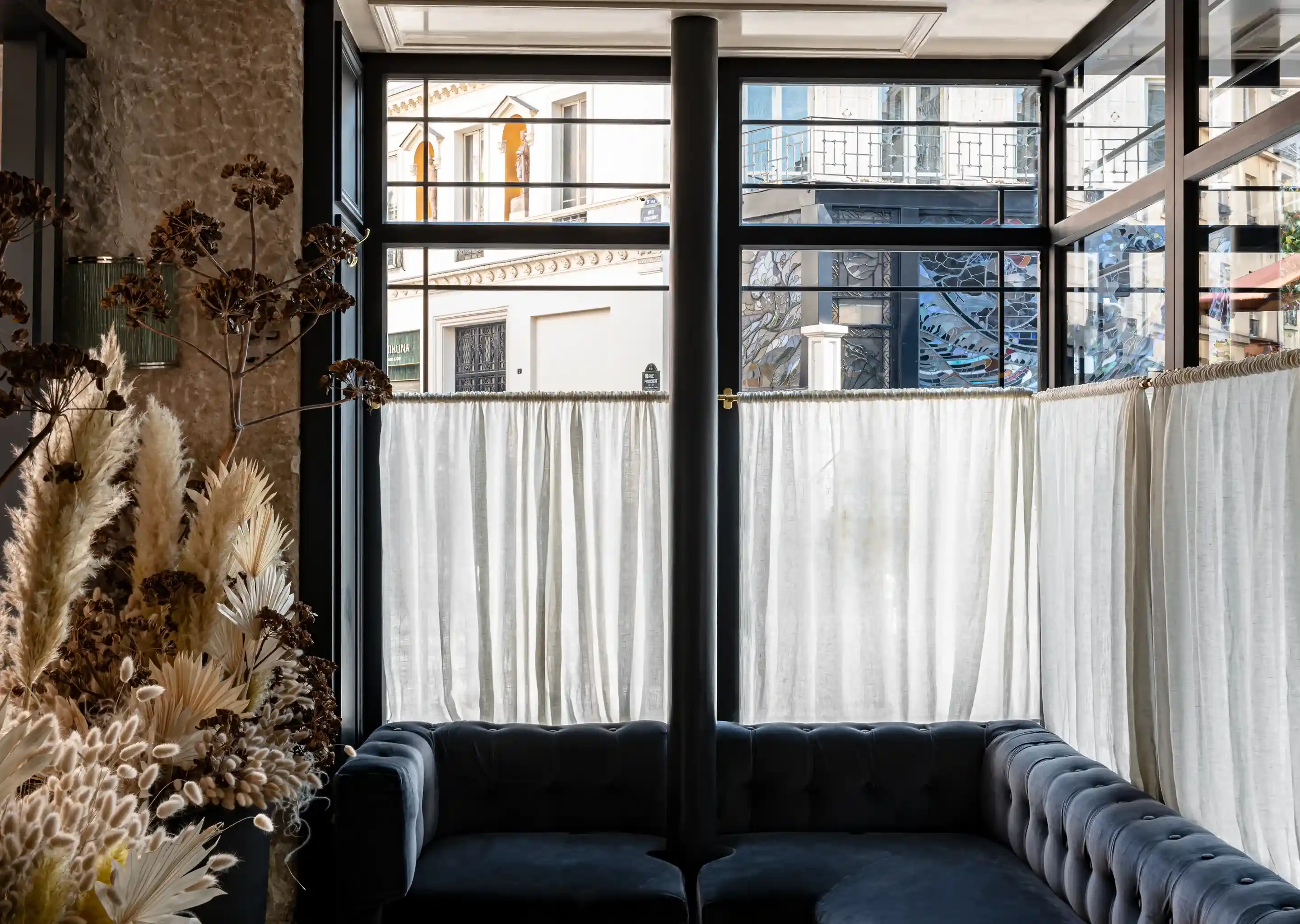 Recanto aconchegante com sofás tufados escuros, arranjo de flores secas e janelas com cortinas de luz em um quarto elegante.