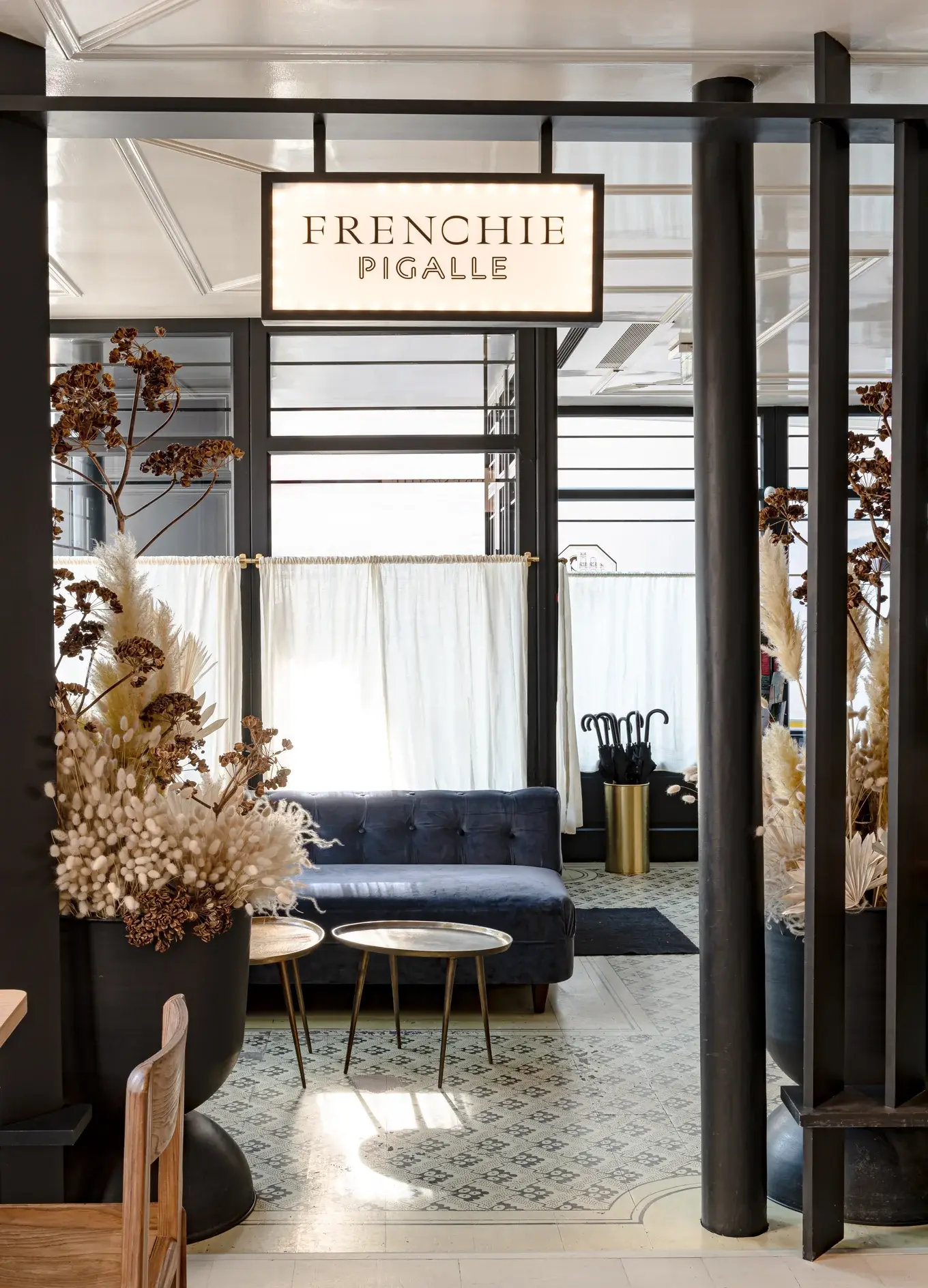 Lounge moderno com sofá de veludo azul, mesas de latão, arranjos florais secos e uma placa Frenchie Pigalle.