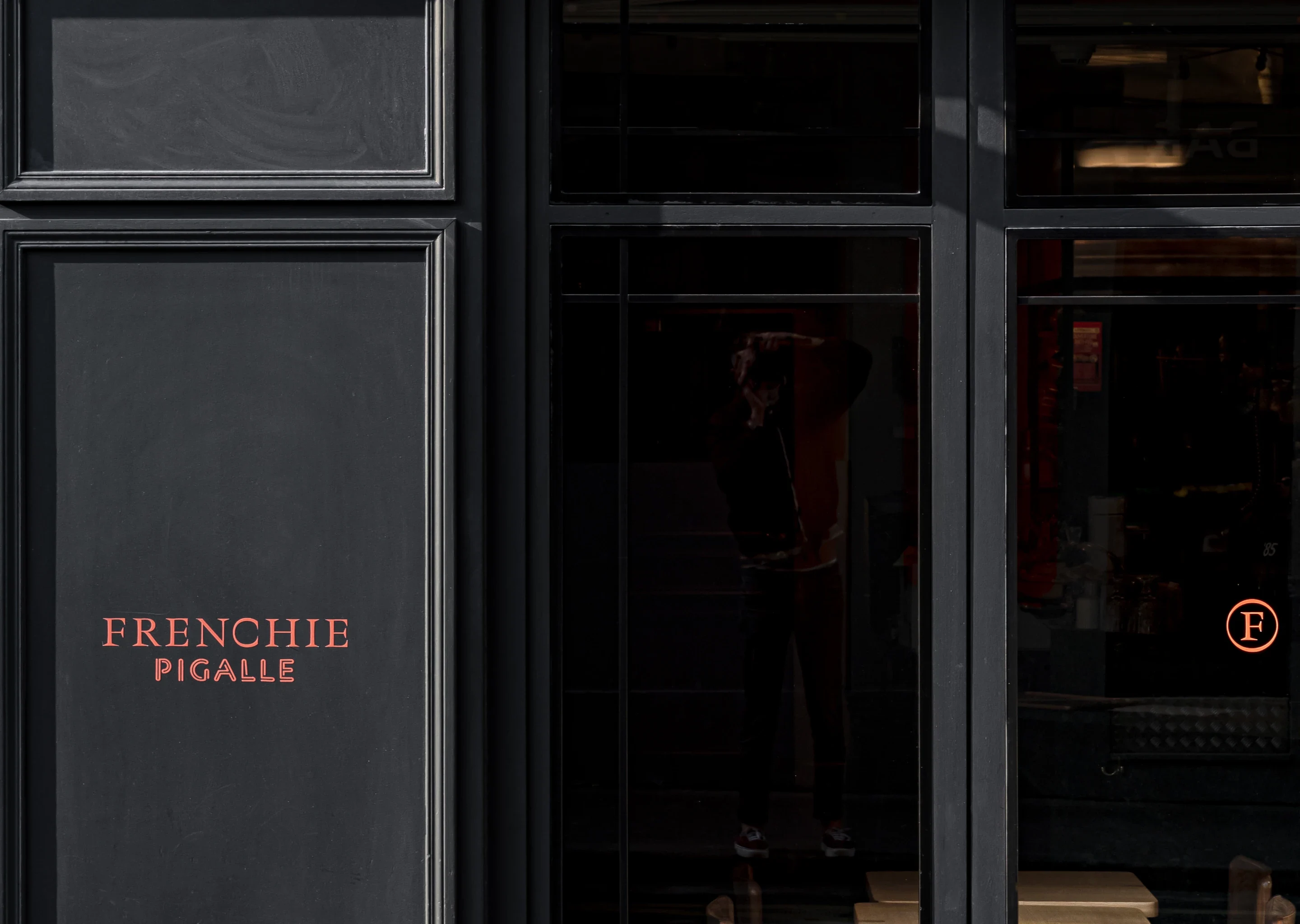 Parede preta com texto vermelho onde se lê “Frenchie Pigalle' ao lado de uma porta de vidro refletindo uma pessoa.