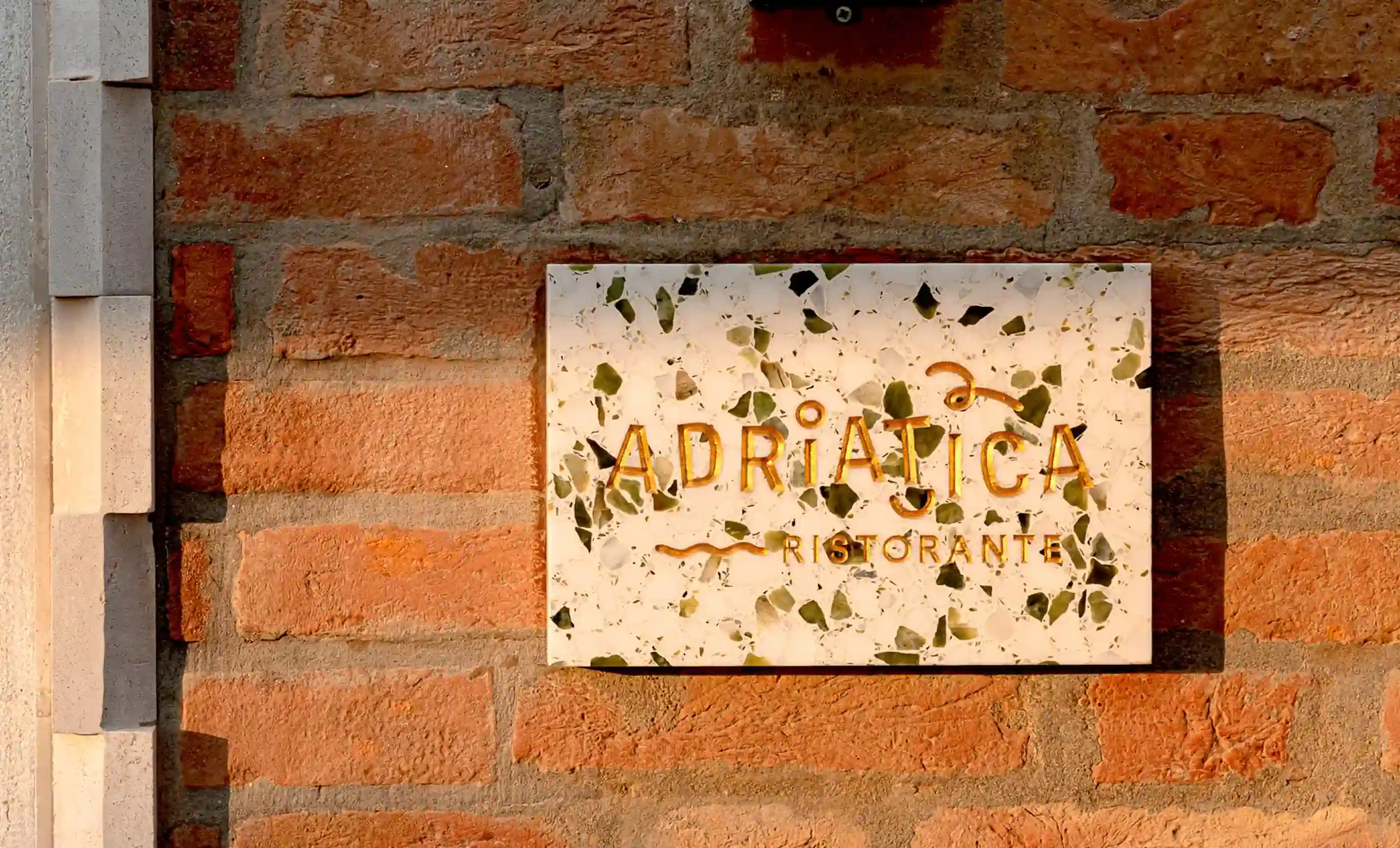 Placa de mosaico montada na parede onde se lê “ADRIATICA Ristorante” em uma parede de tijolos.