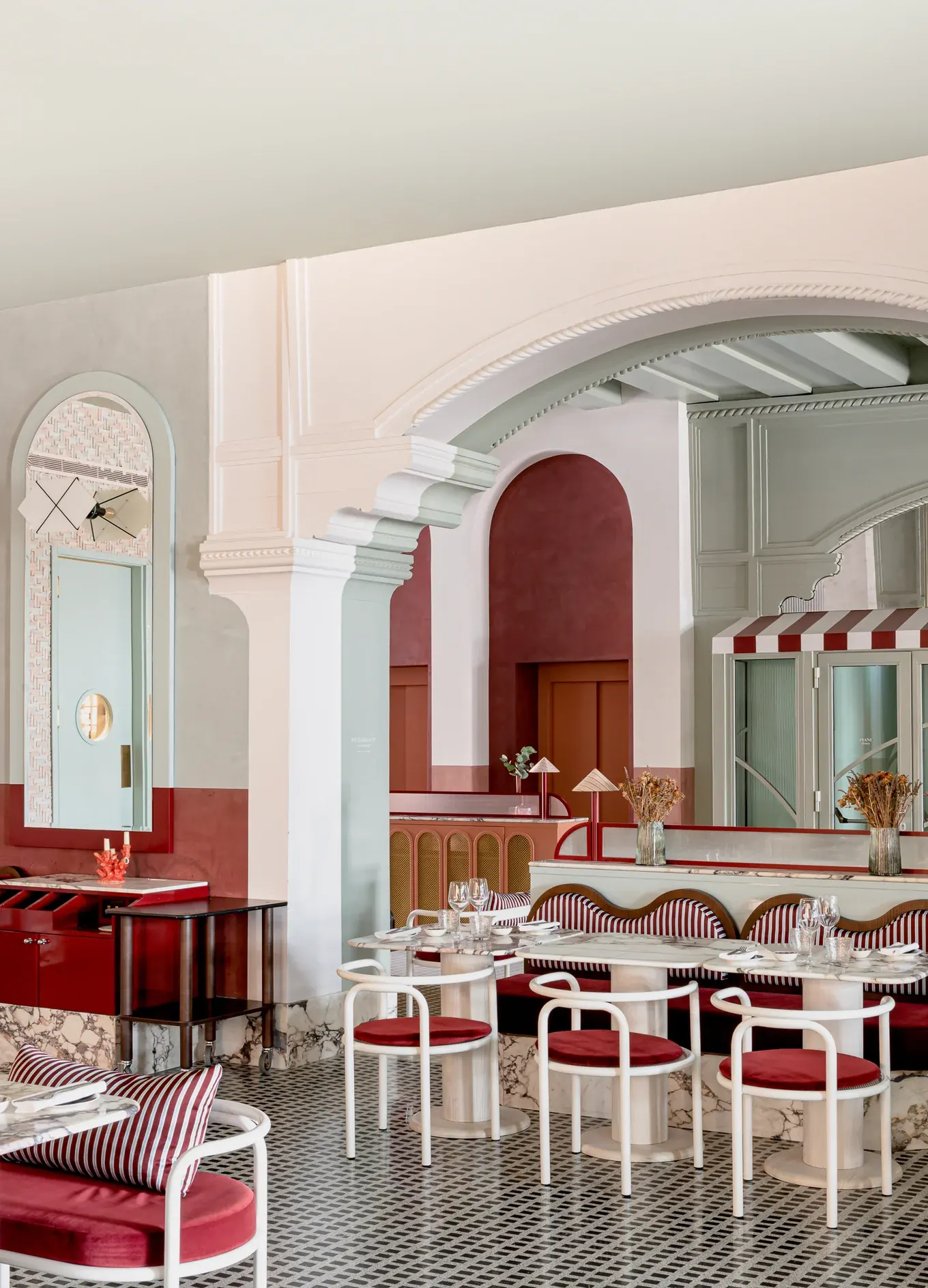 Área de jantar interna com cadeiras brancas e vermelhas, mesas de mármore, portas em arco e espelhos decorativos.