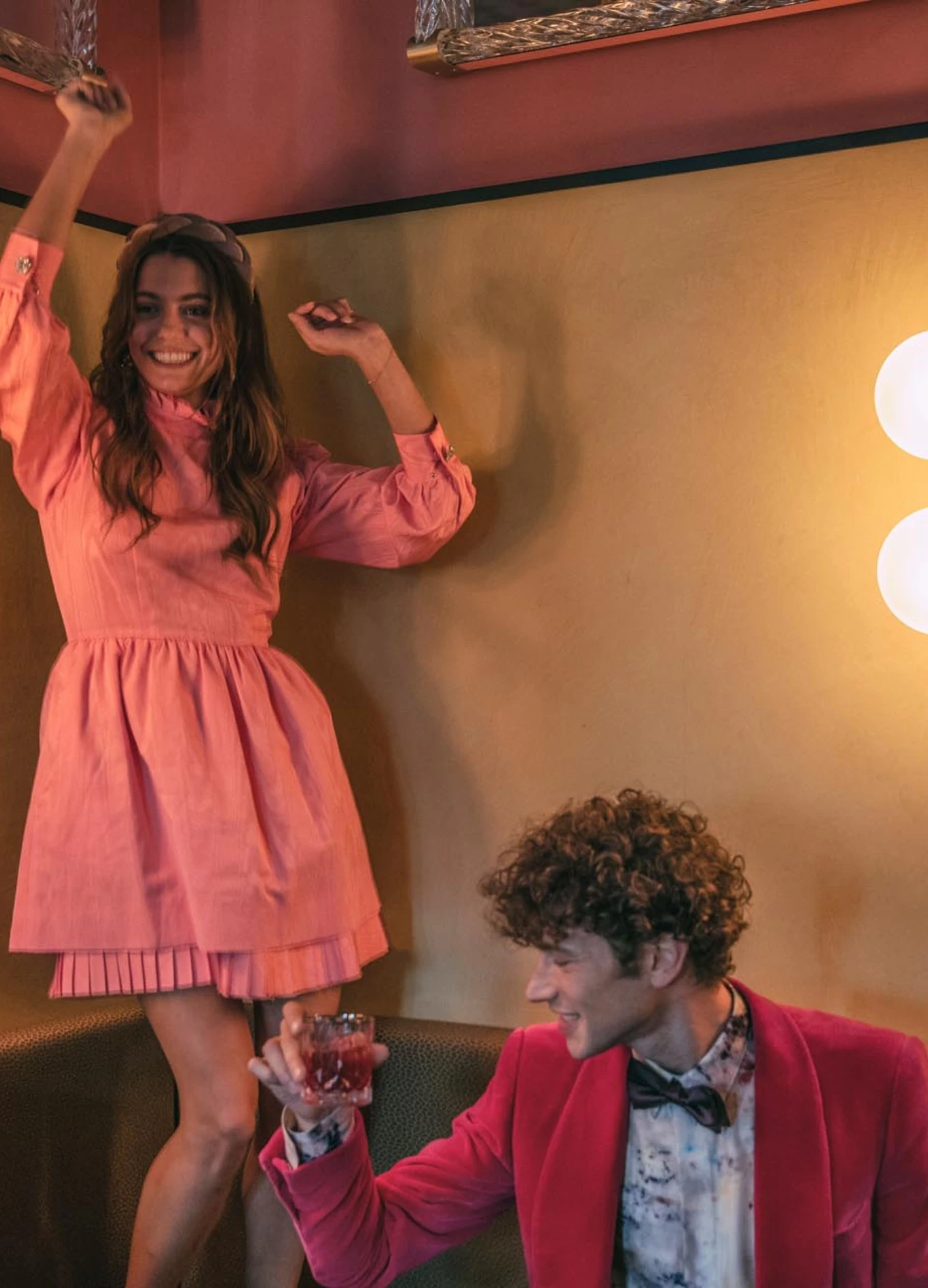 Mulher de vestido rosa dançando ao lado de um homem de jaqueta vermelha segurando uma bebida em um quarto aconchegante.