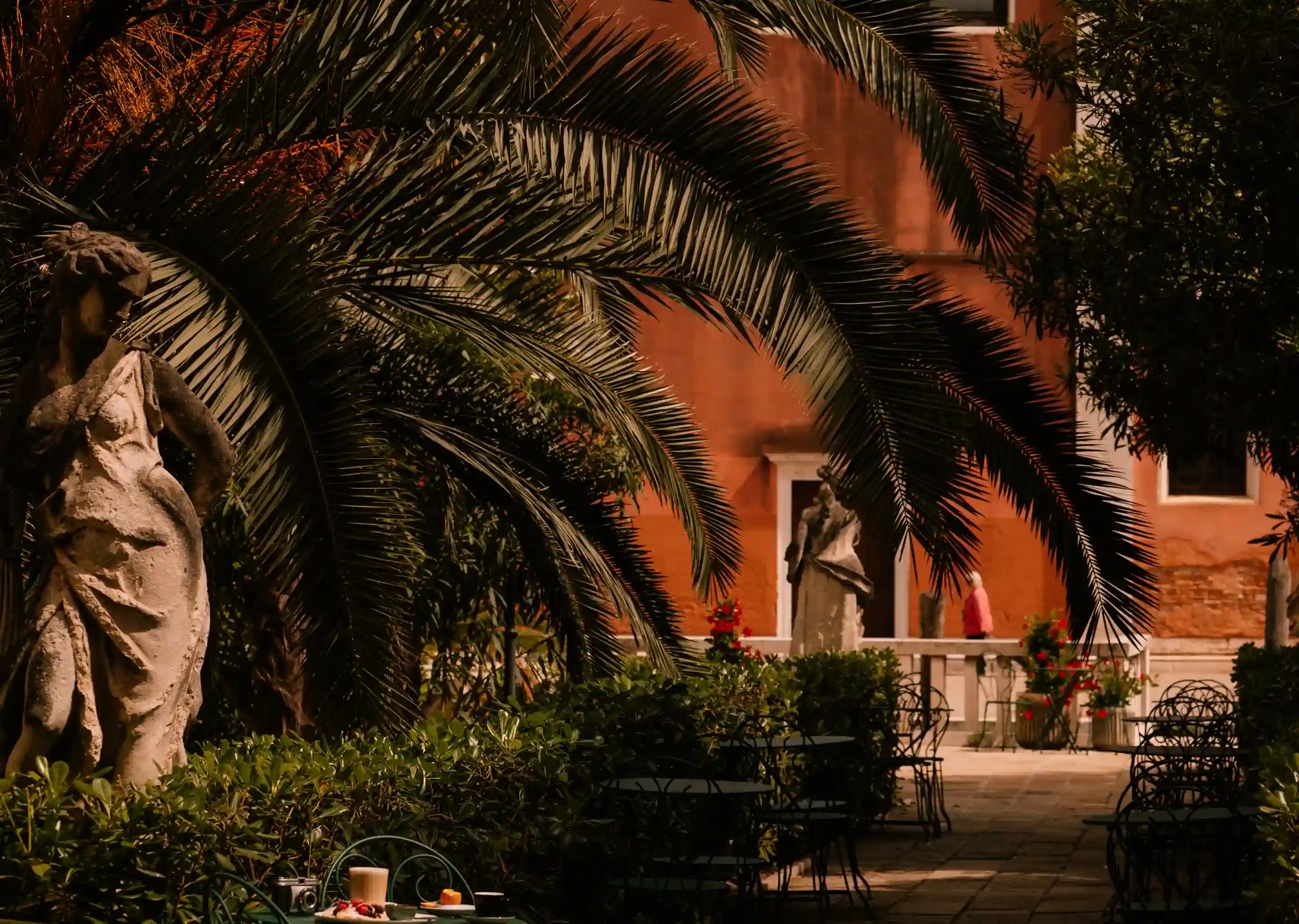 Jardim ao ar livre com estátuas de pedra, folhas verdes de palmeira, mesas de ferro forjado e uma bebida na mesa.