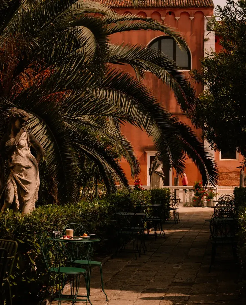 Mesas ao ar livre com cadeiras verdes e mesa sob grandes folhas de palmeira ao lado de estátuas e prédio laranja.