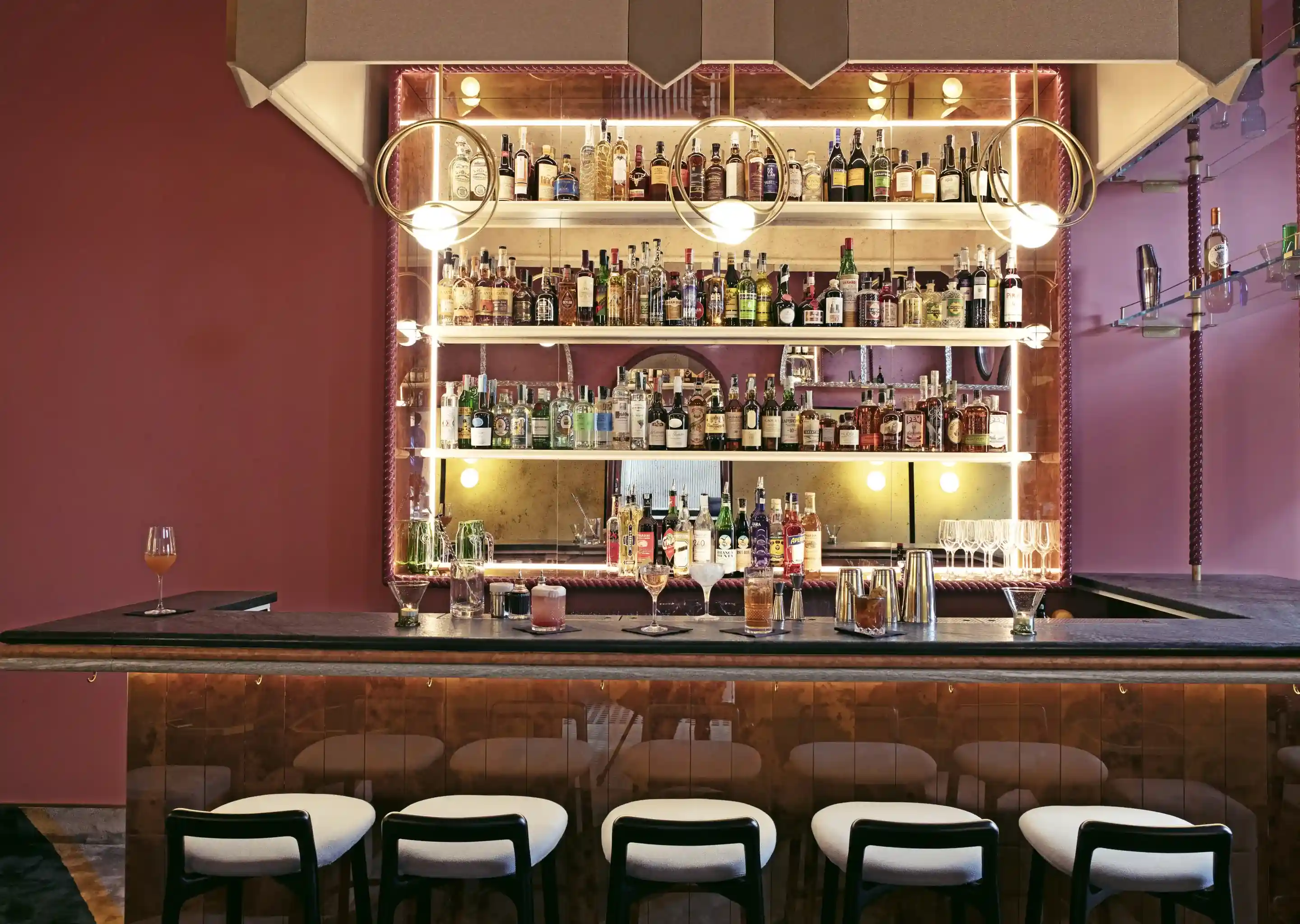 Bar de coquetéis moderno com prateleiras de bebidas iluminadas, luzes penduradas, seis bancos almofadados e bebidas no balcão.