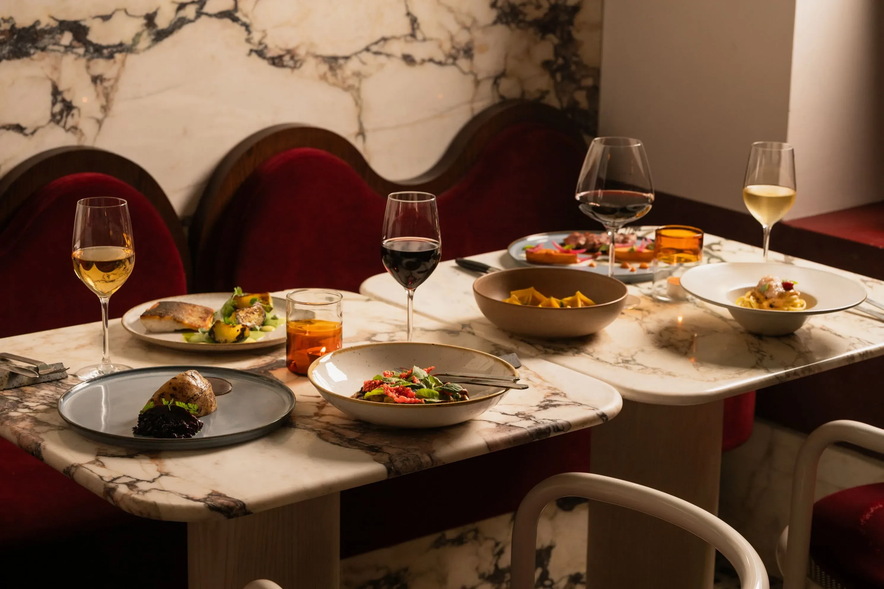 Mesa de mármore com pratos elegantes, taças de vinho e assentos de veludo vermelho em um ambiente requintado.