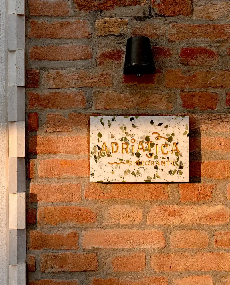 Placa de terrazzo branco com texto dourado lendo Adriatica Ristorante em uma parede de tijolos vermelhos sob uma luminária preta.