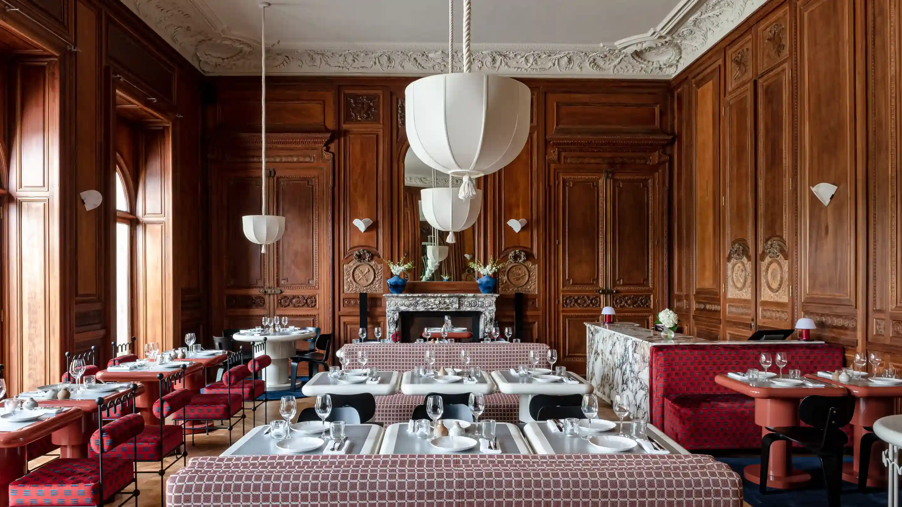 Ristorante elegante con pareti rivestite in legno, sedute rosse a motivi geometrici, tavoli bianchi e grandi lampade a sospensione.