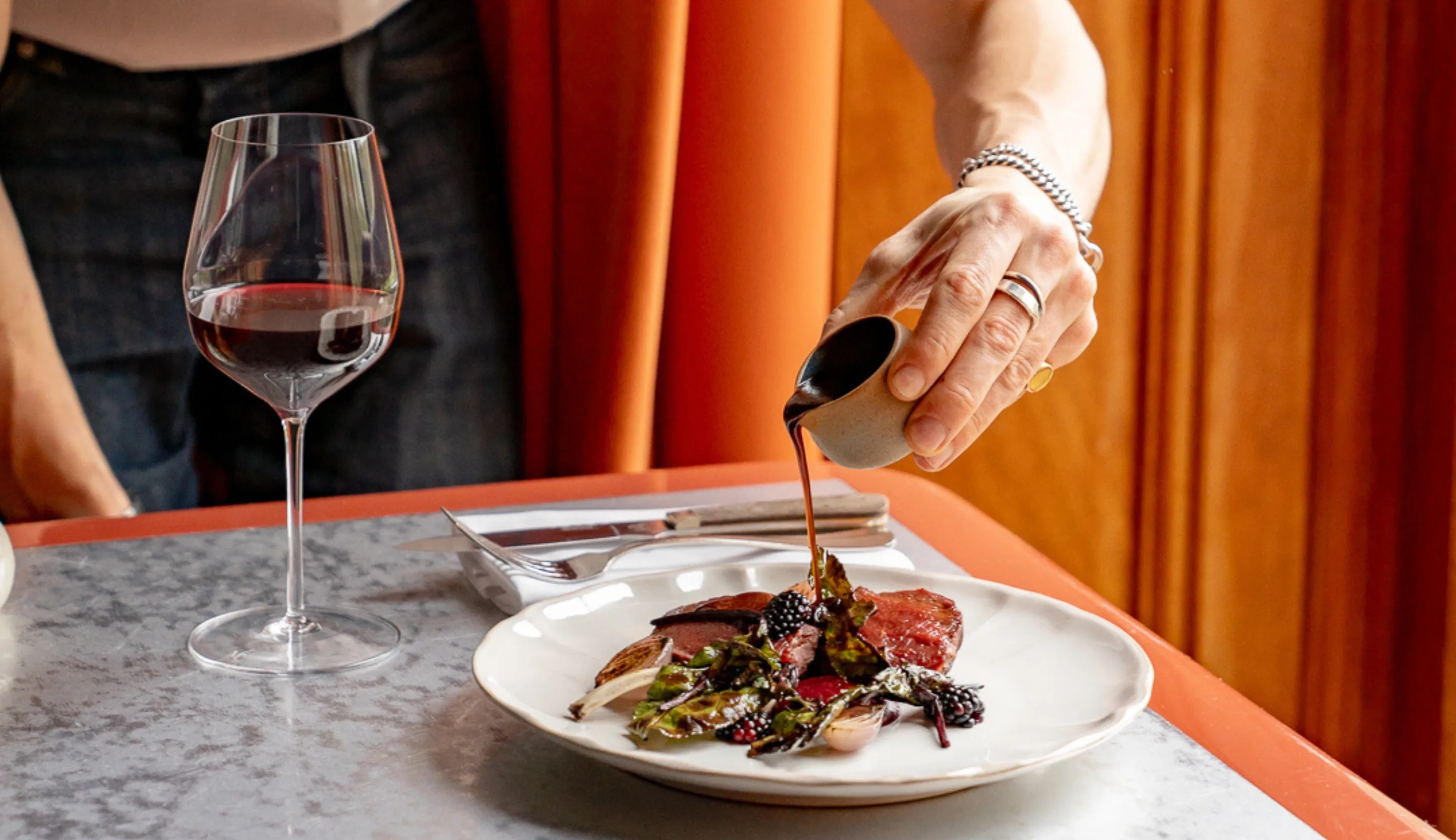 Versare a mano la salsa su carne gourmet placcata con verdure e more accanto a un bicchiere di vino rosso.