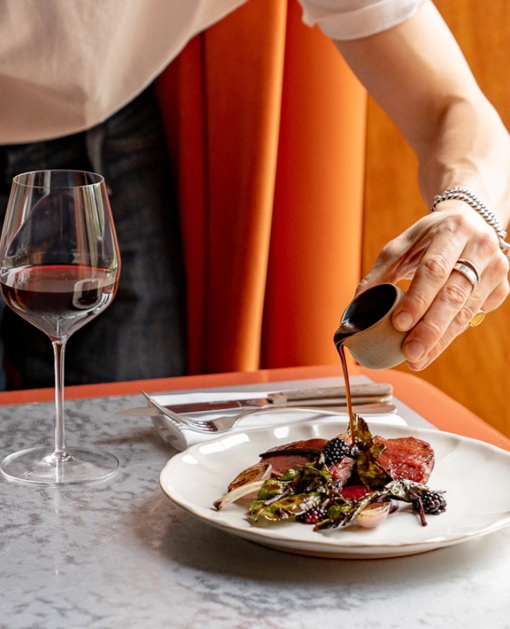 Persona che versa la salsa su un piatto gourmet con verdure e frutti di bosco accanto a un bicchiere di vino rosso.