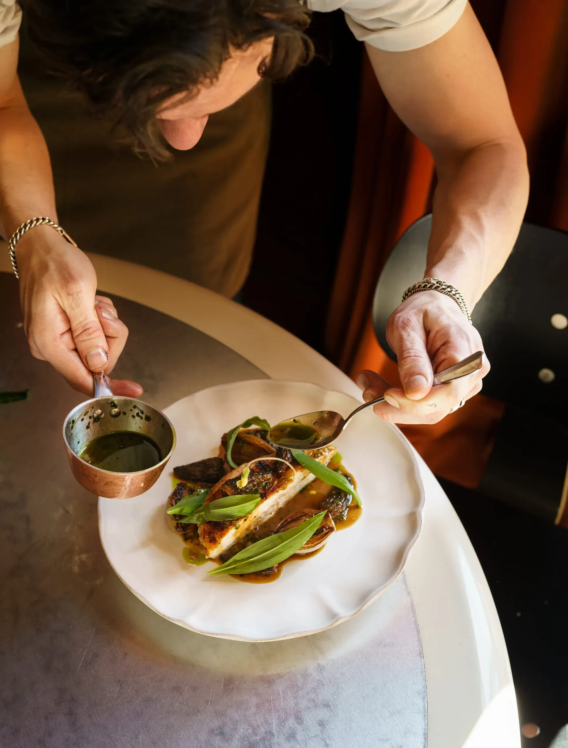 Cuoco che indossa braccialetti che spruzzano salsa verde su pesce placcato guarnito con foglie e verdure.