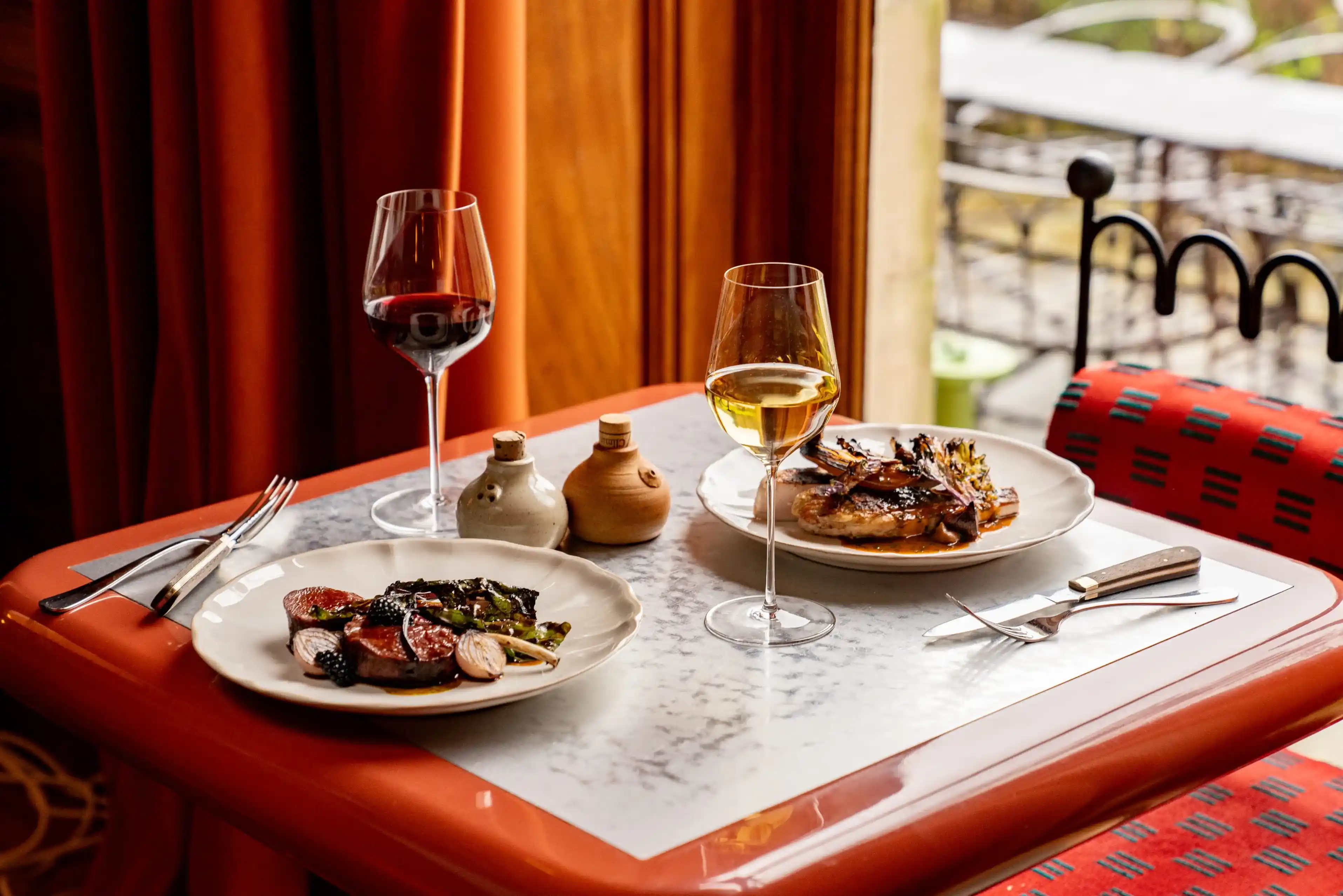Tavolo con due piatti di cucina gourmet, bicchieri da vino rosso e bianco e saliera e pepiera.
