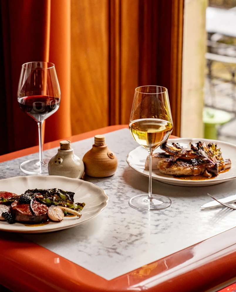 Tavolo con due piatti di cucina gourmet, bicchieri di vino rosso e bianco e saliera e pepiera.