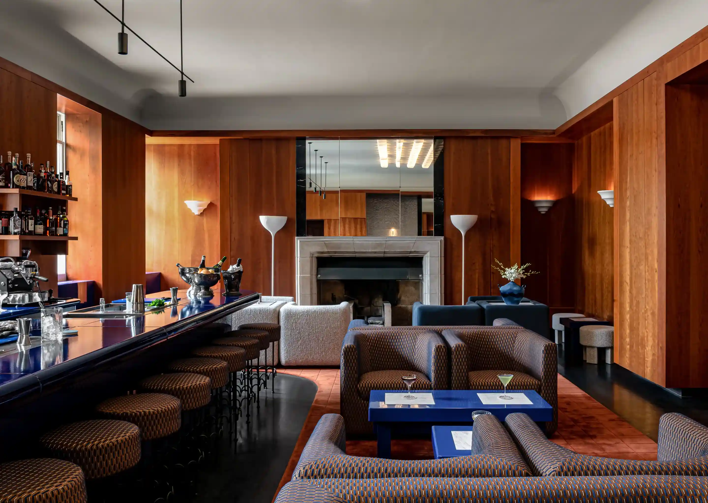 Moderno cocktail bar con sedie a motivi geometrici, tavoli blu, pareti in legno e un camino sullo sfondo.