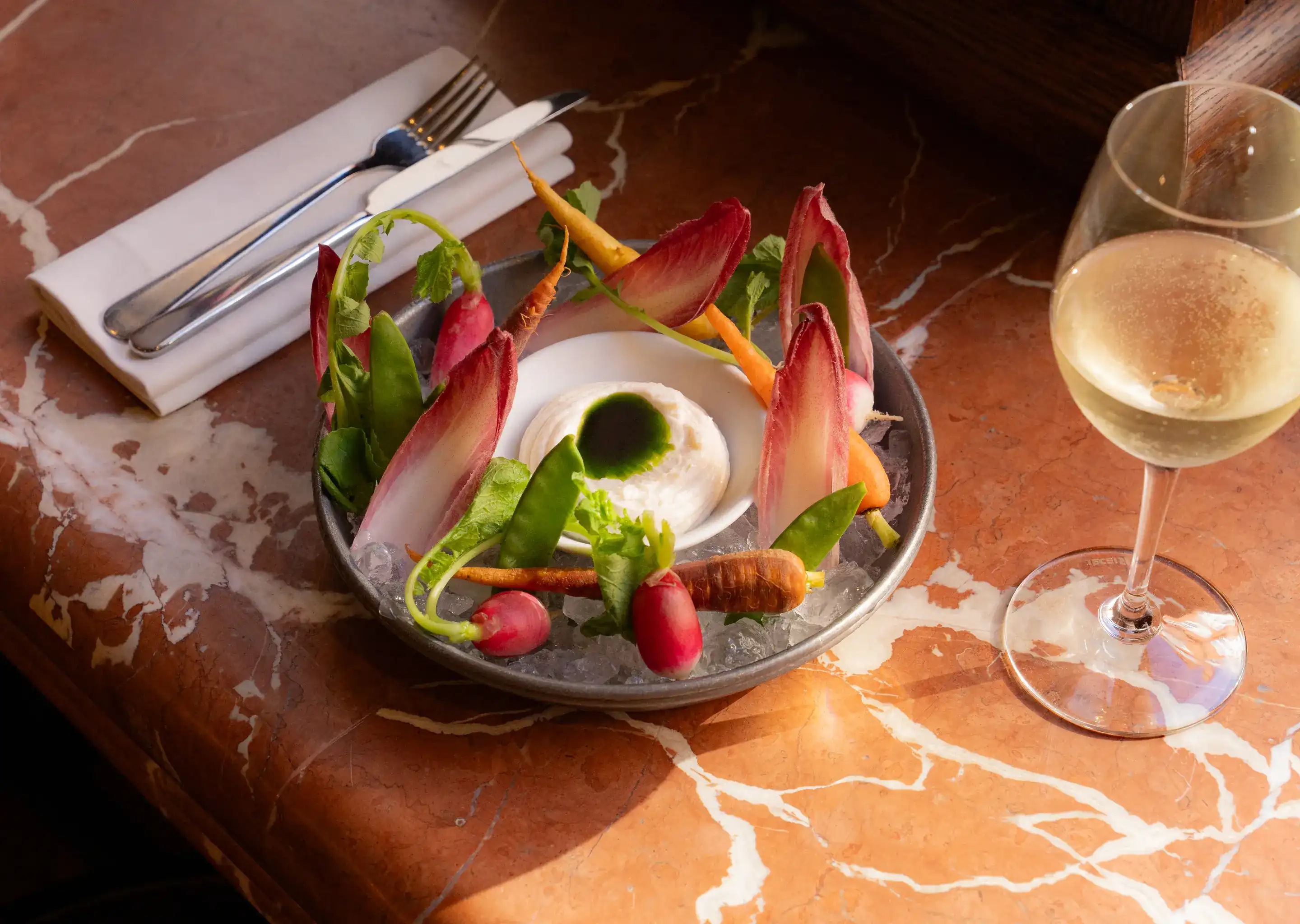 Ciotola di ravanelli freschi, carote e verdure con salsa di crema e un bicchiere di vino bianco sul tavolo di marmo.