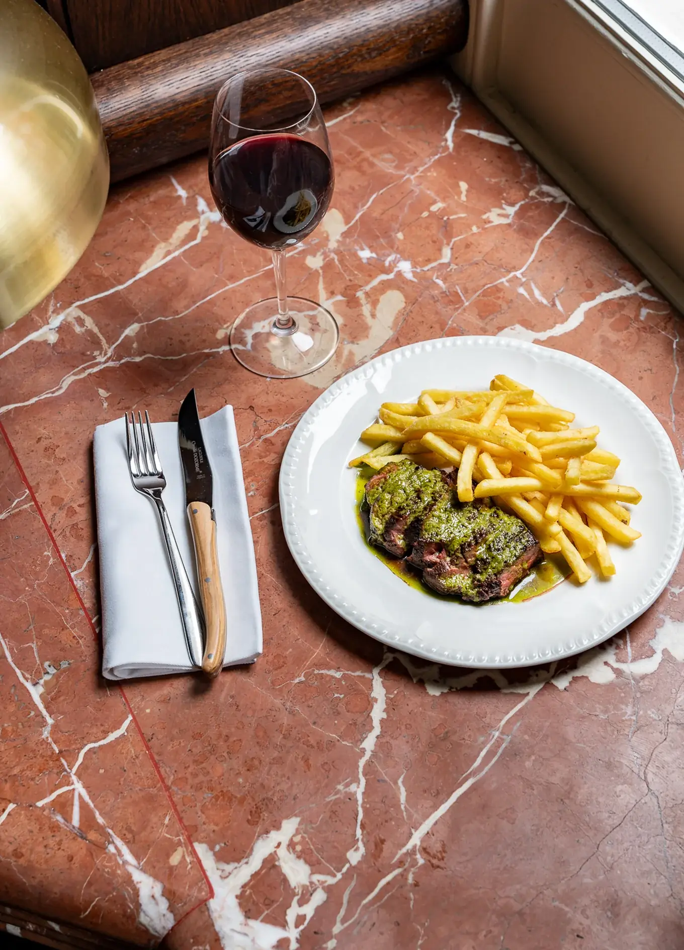 Piatto con bistecca condita con salsa verde e patatine fritte su un tavolo di marmo con vino e posate.