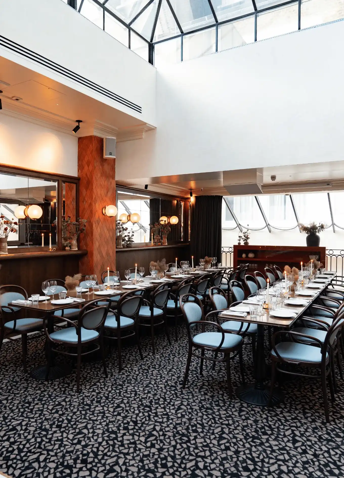 Tavoli da pranzo lunghi con sedie blu e candele in un luminoso ristorante con soffitto a lucernario.