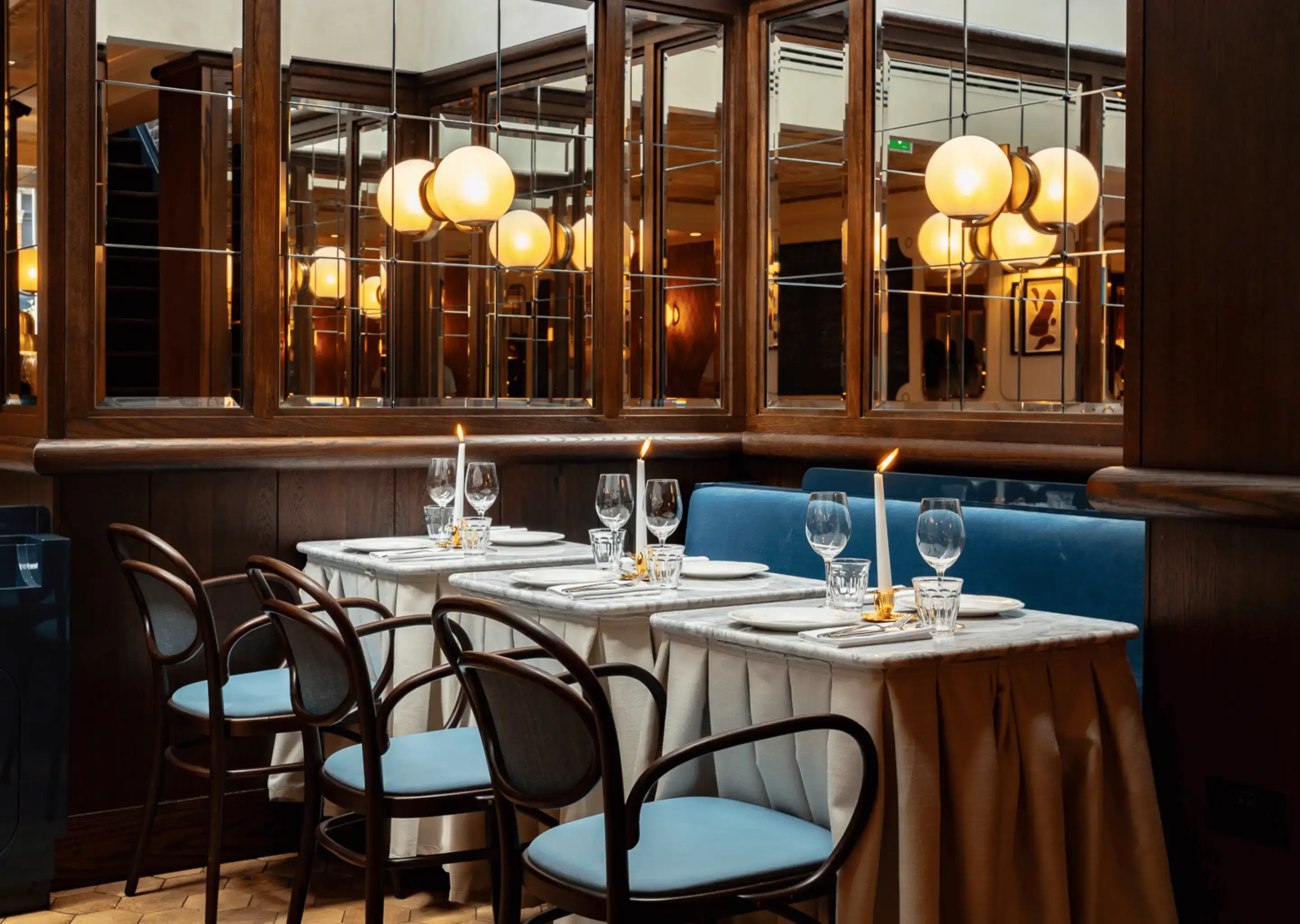 Elegante angolo ristorante con pannelli in legno, specchi, sedie blu, lenzuola bianche e candele accese sui tavoli.