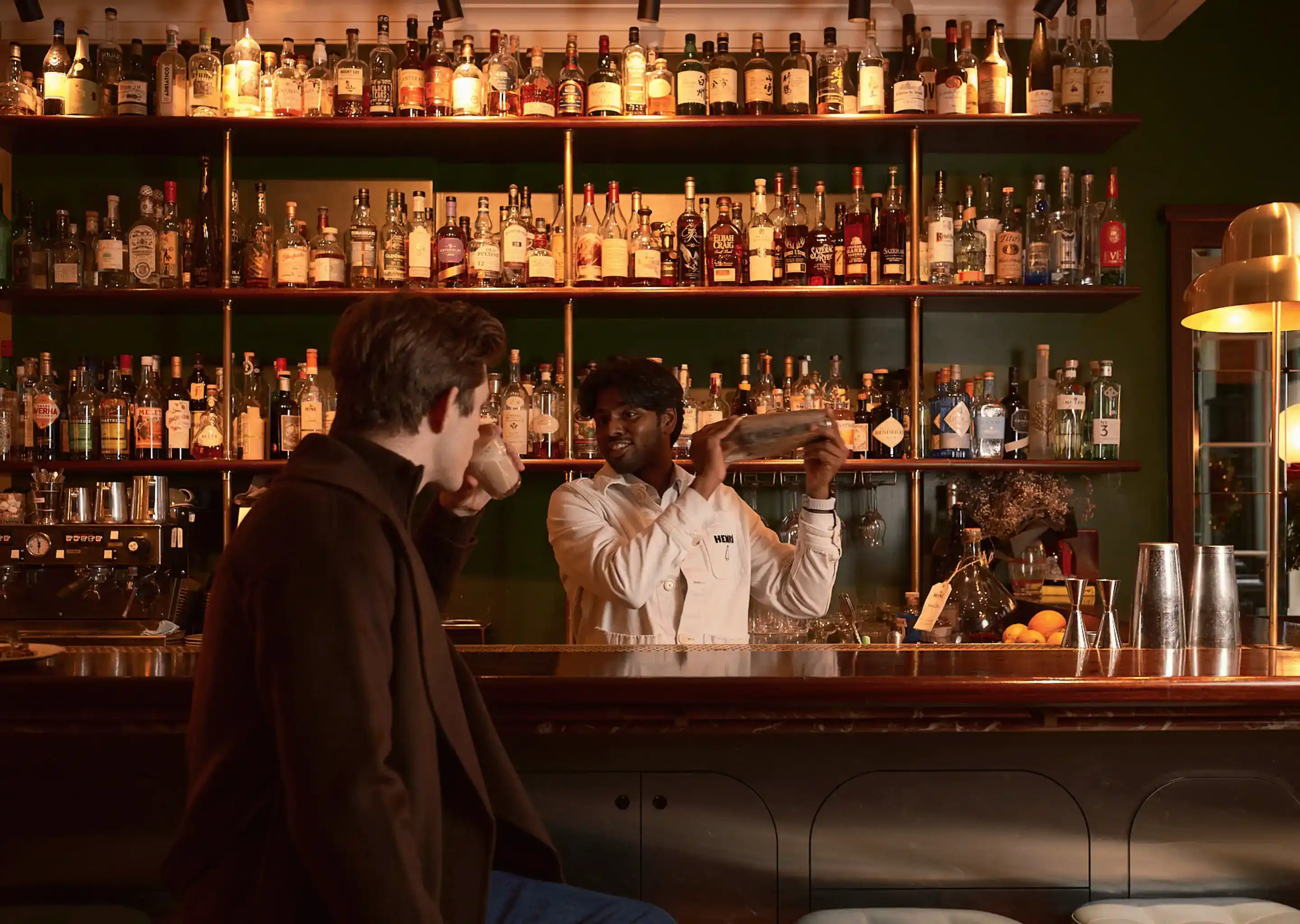 Barista che scuote un cocktail dietro un bar mentre un uomo con un cappotto marrone beve una bevanda.