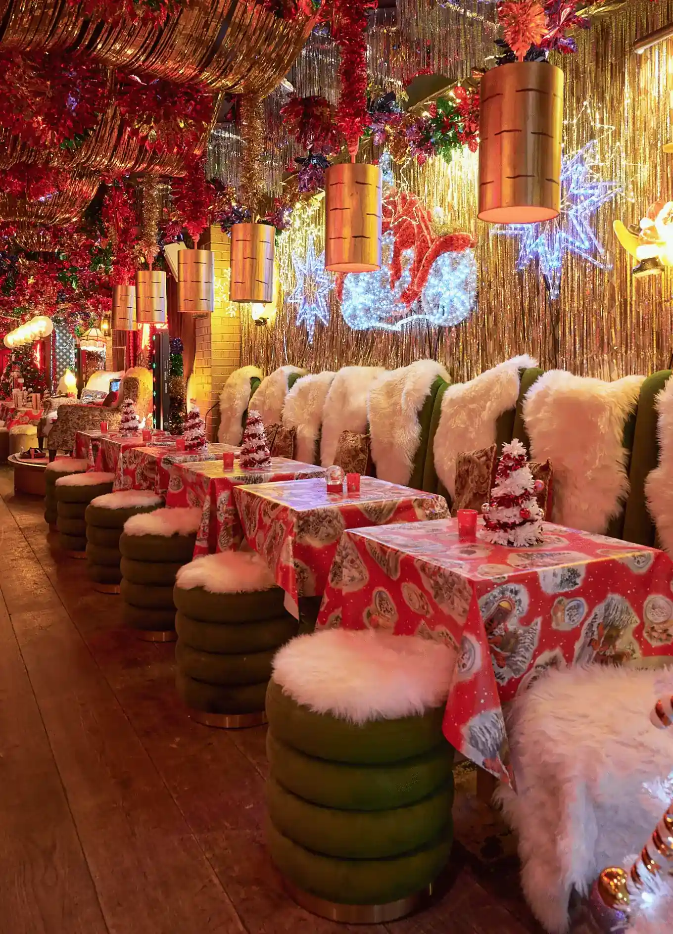 Assentos de restaurante com tema natalino com toalhas de mesa vermelhas, almofadas de pele, luzes penduradas e decorações festivas.