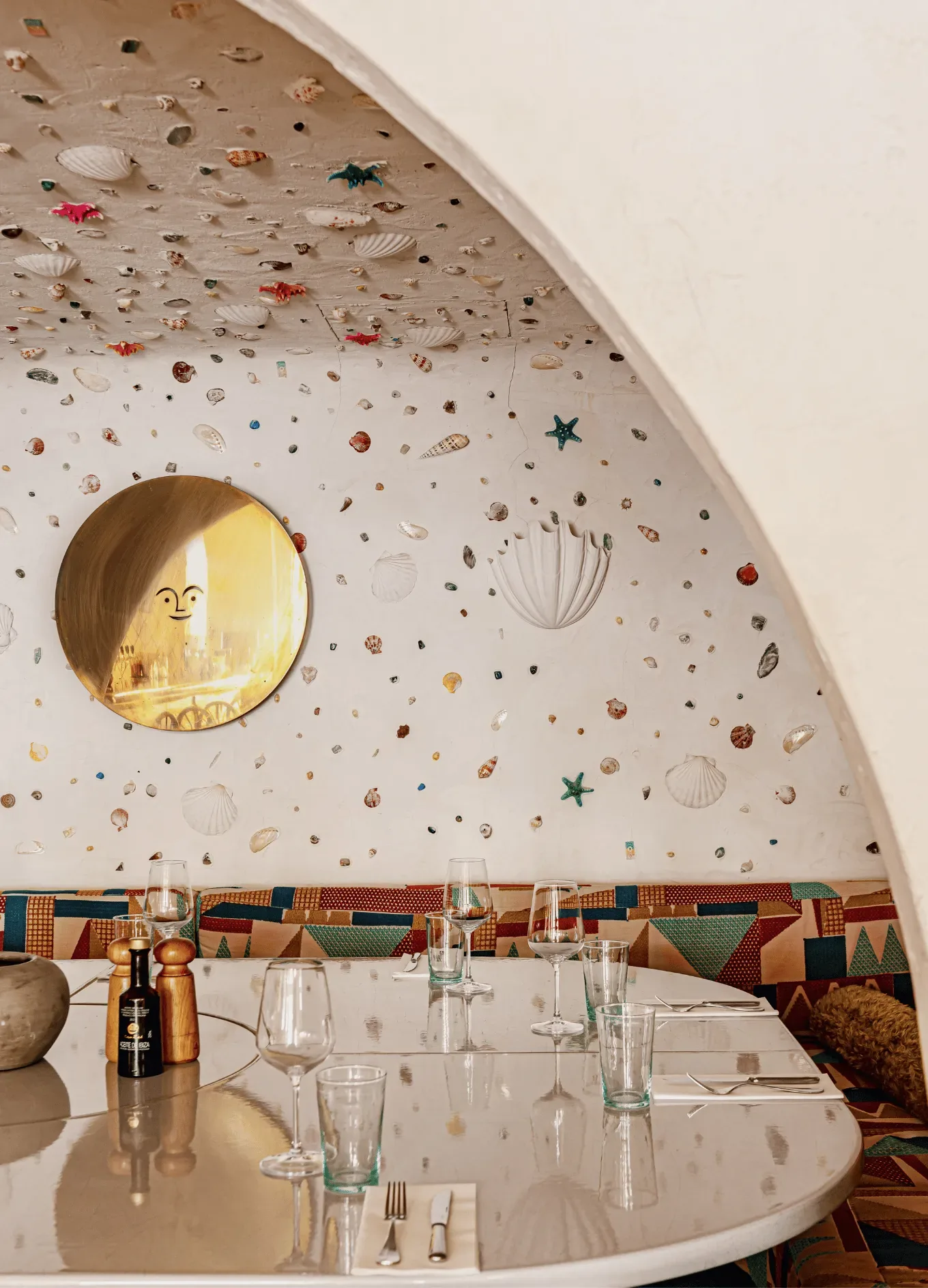 Sala da pranzo curva con parete decorata con conchiglie bianche, sedute colorate, specchio rotondo e tavolo apparecchiato.