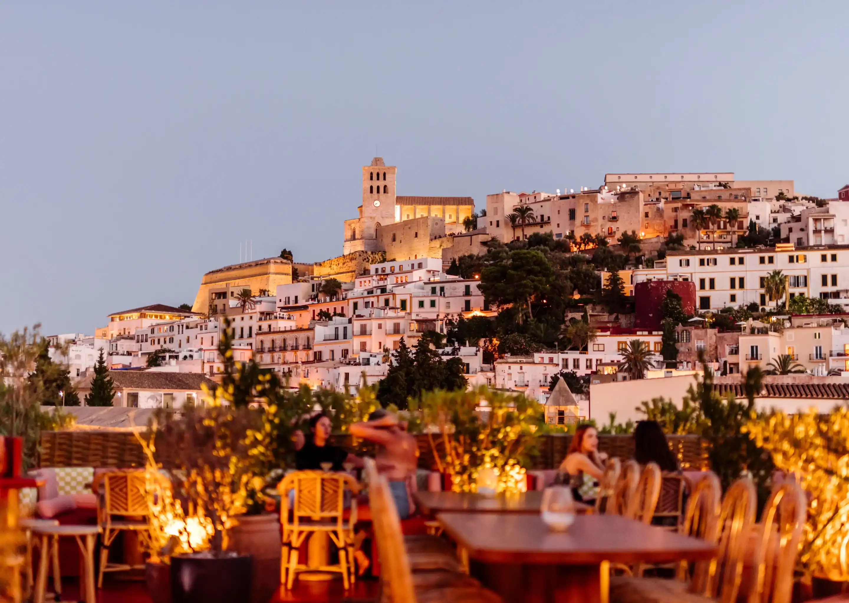 Cena serale sul tetto con gente e luci calde con vista su una città collinare con un campanile.