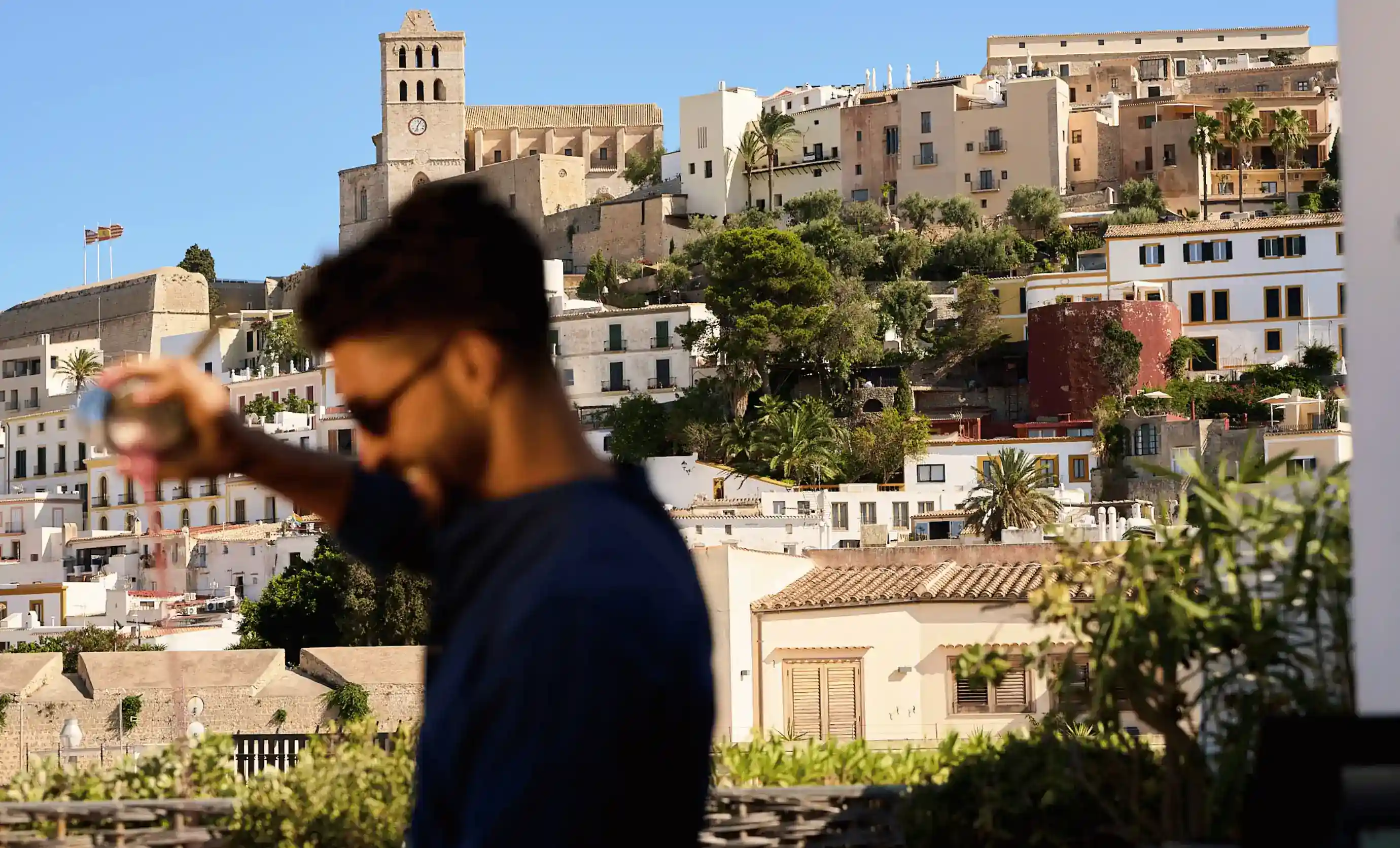 Uomo in occhiali da sole che versa liquido da uno shaker con la storica città collinare e la torre dell'orologio sullo sfondo.