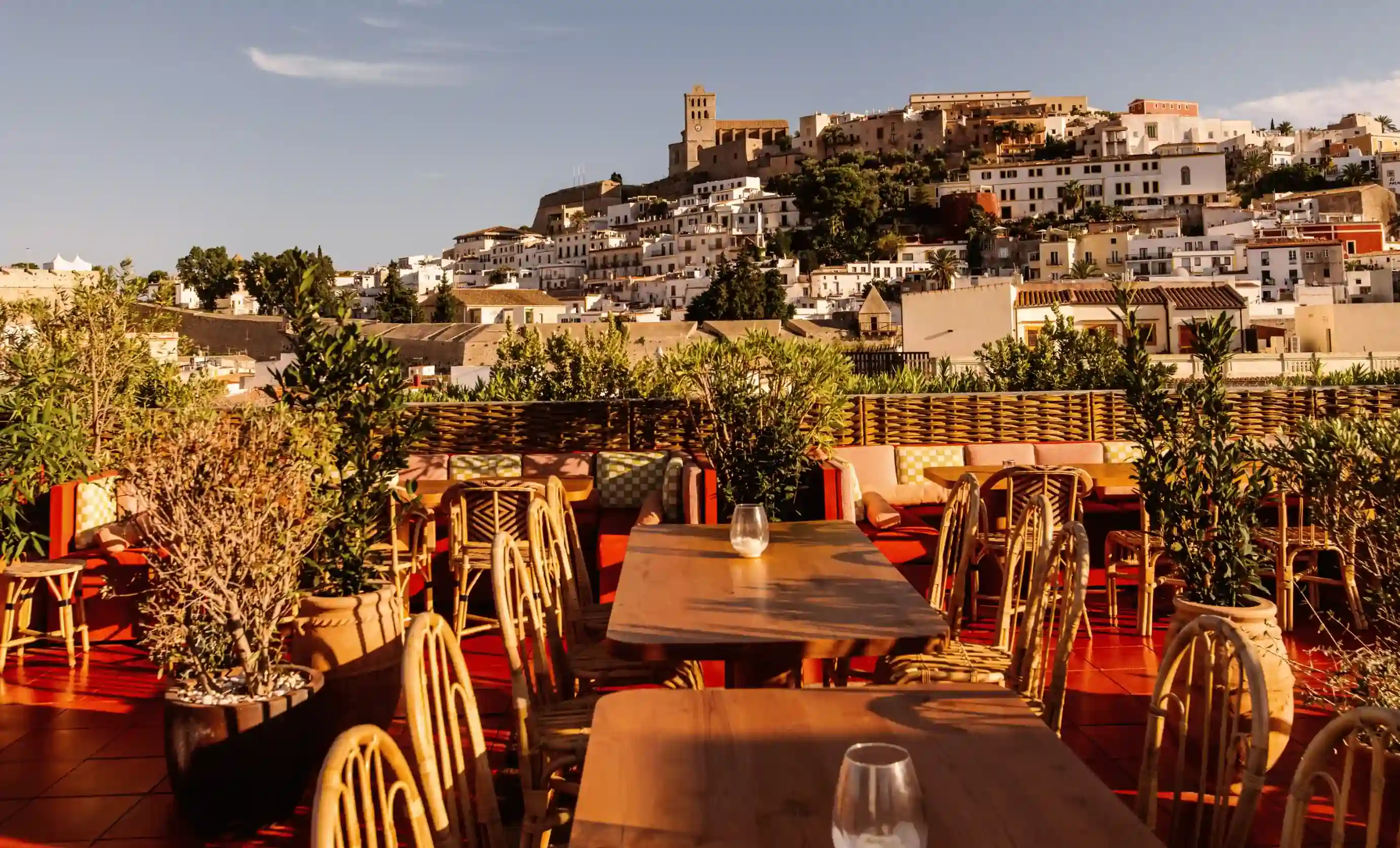 Terrazza all'aperto con tavoli in legno, sedie in vimini, piante e città collinare sullo sfondo sotto un cielo limpido.