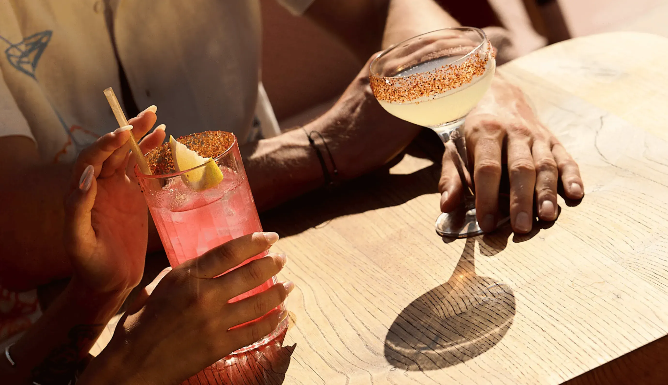 Due persone che tengono cocktail con bicchieri bordati di peperoncino e spicchi di limone su un tavolo di legno.