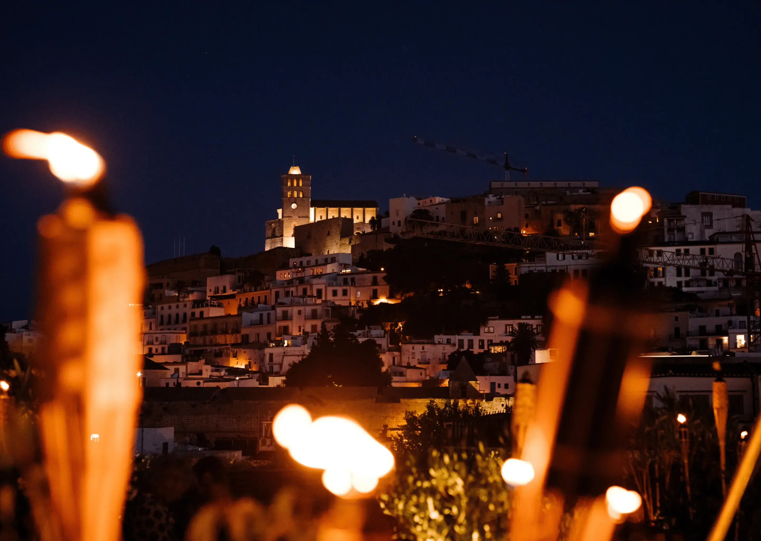 Vista notturna di una città collinare con chiesa illuminata e fiamme di torce offuscate in primo piano.