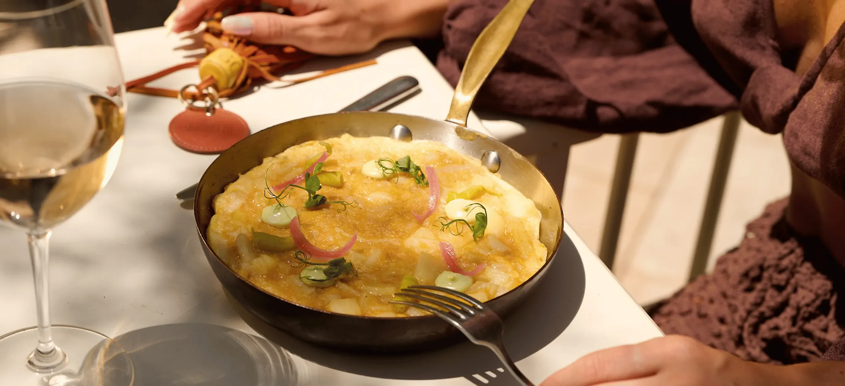 Padella con frittata guarnita con erbe aromatiche, cipolle in salamoia e salsa su un tavolo bianco vicino a un bicchiere di vino.