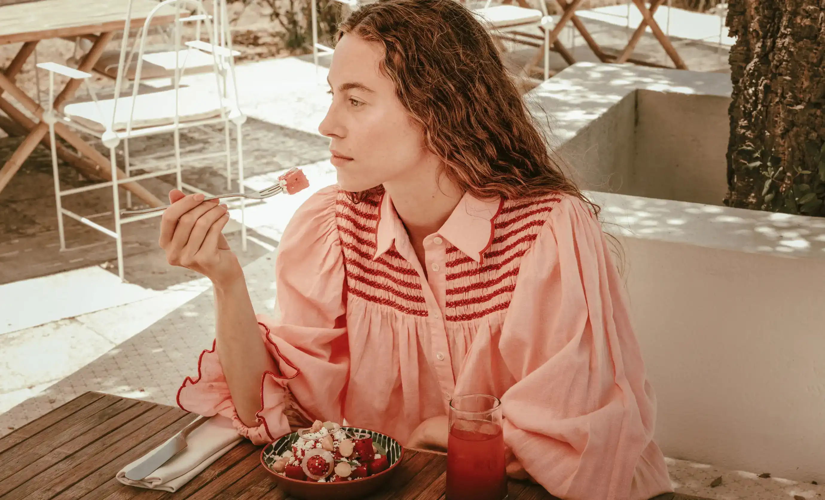Donna in camicetta rosa che mangia un'insalata con anguria e beve una bevanda rossa all'aperto.