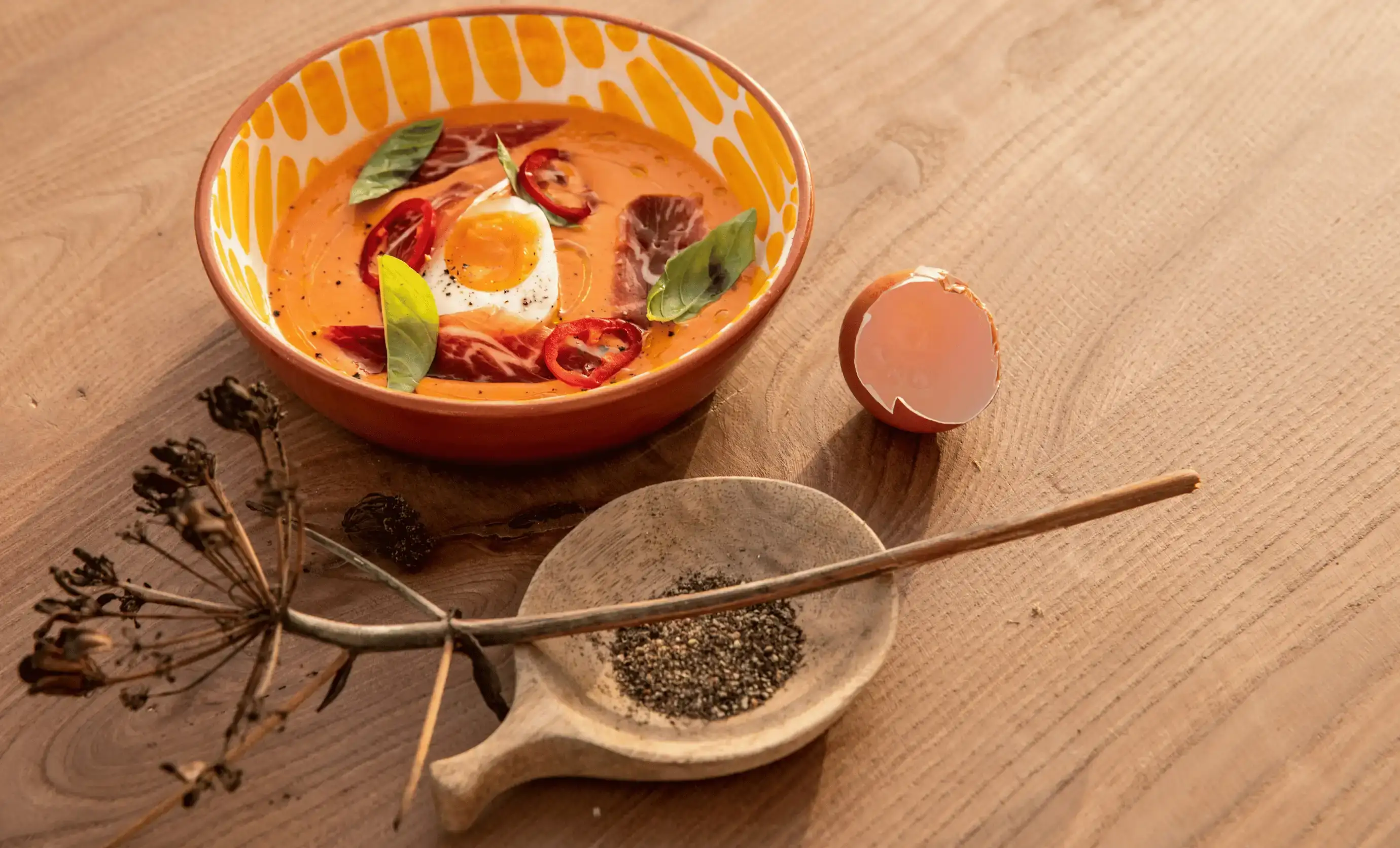 Ciotola con zuppa d'arancia, uovo sodo, foglie di basilico, fette di peperoncino, accanto a guscio d'uovo e pepe sul tavolo di legno.