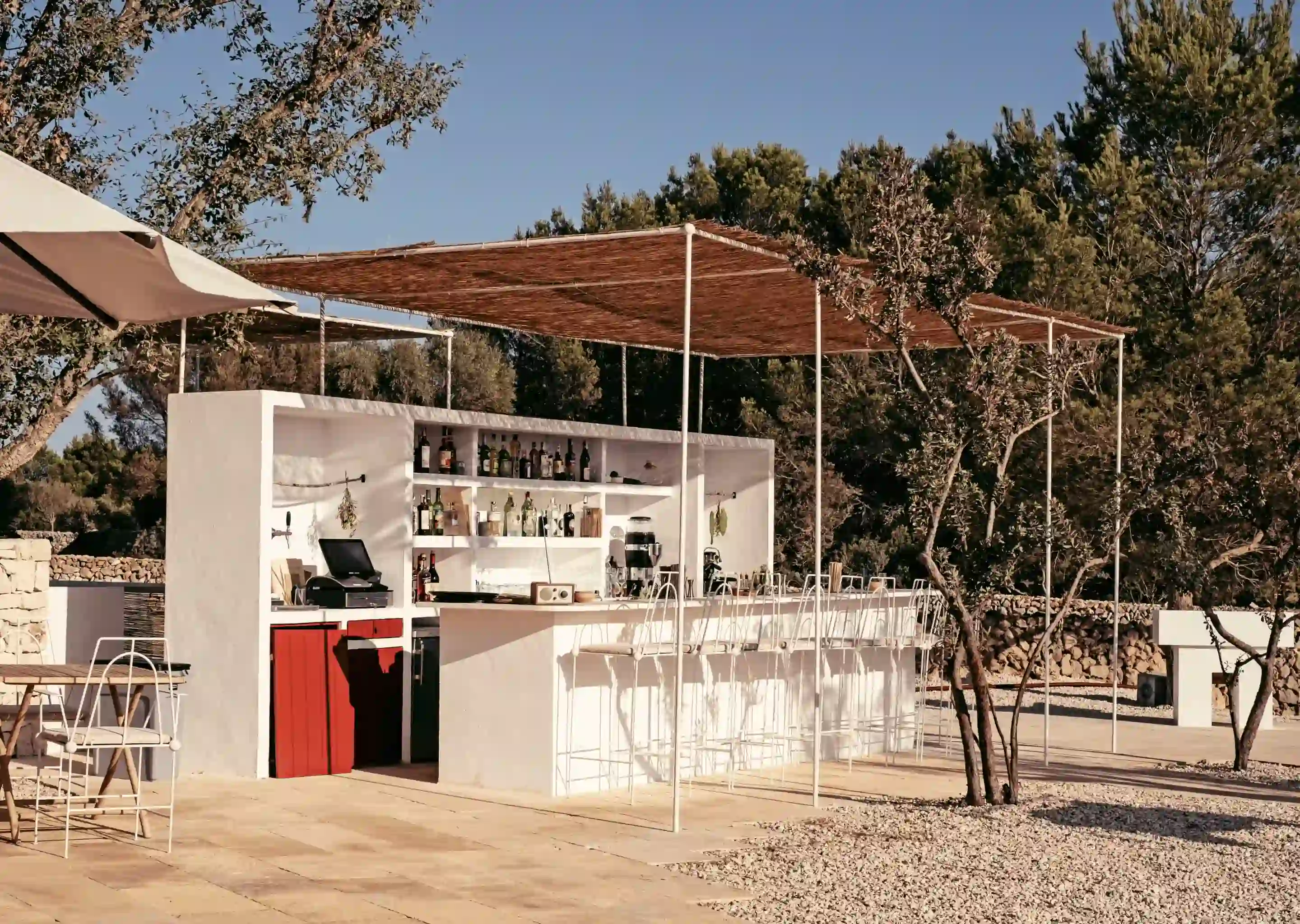 Bar all'aperto dell'hotel con bancone bianco, sgabelli alti, ombreggiato da un pergolato in legno e circondato da alberi.